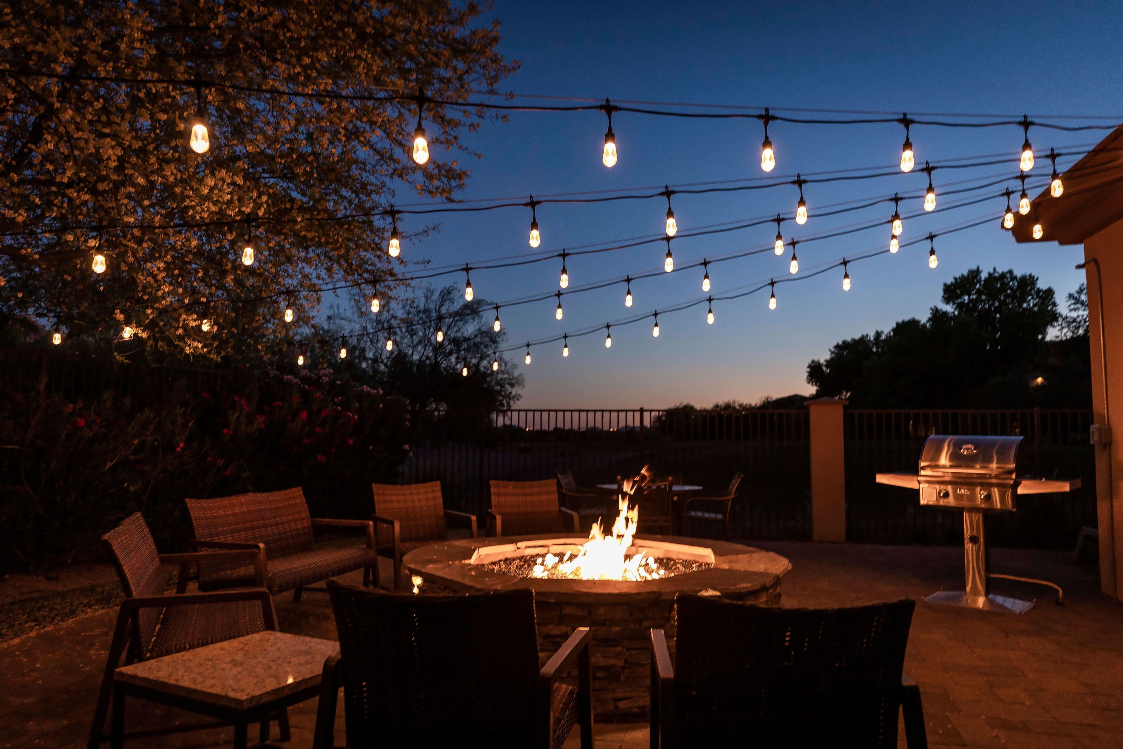 An outdoor seating area with a fire pit, surrounded by chairs and tables, under string lights.