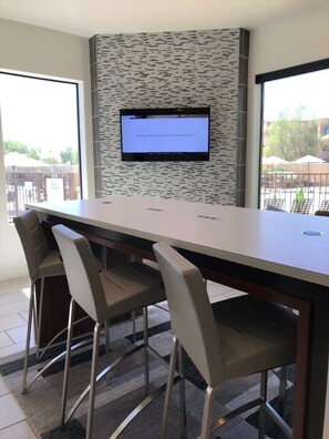Un salón de conferencias con una larga mesa, cuatro sillas, un televisor de pantalla plana montado en una pared texturizada y grandes ventanales con vistas a una zona de estar exterior.