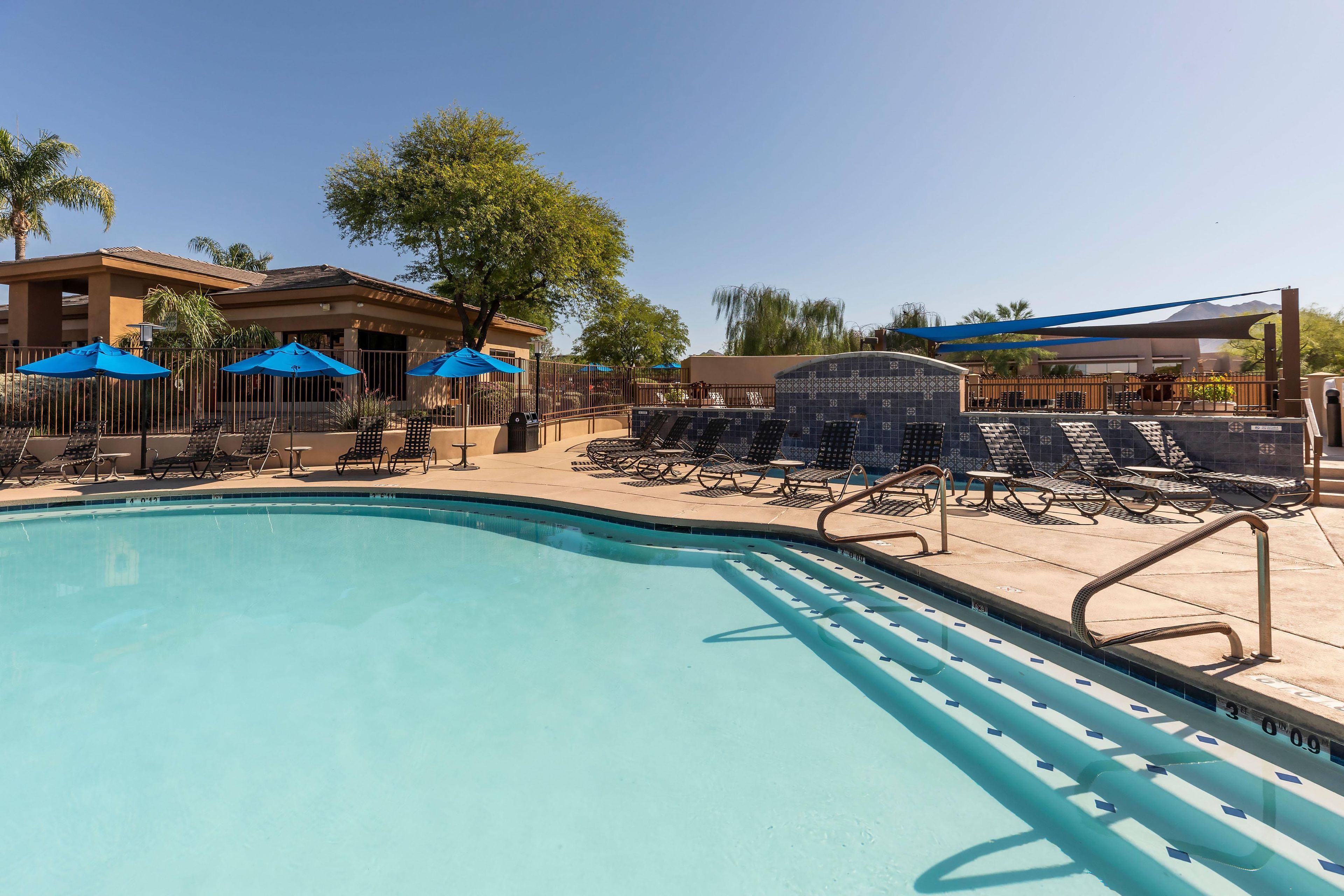 A swimming pool with lounge chairs and umbrellas, surrounded by a fence and trees.