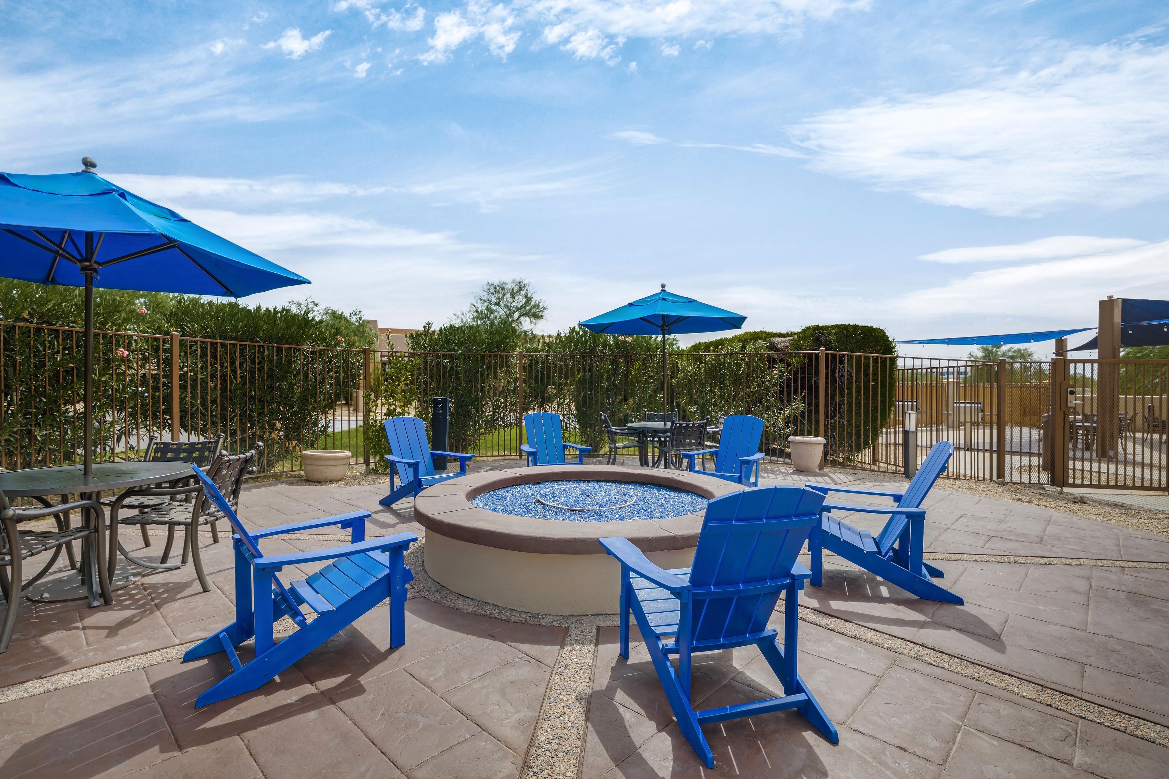 An outdoor patio area with a circular fire pit, blue chairs, and umbrellas.