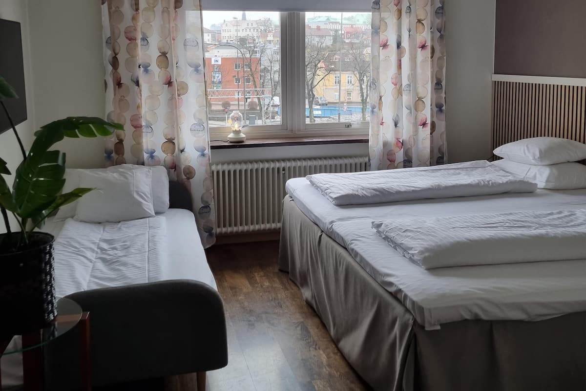 Habitación de hotel con dos camas, un radiador, una planta en maceta y vistas a edificios a través del ventanal.