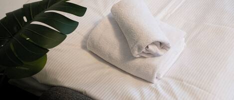 A neatly made bed with a rolled white towel and a large green plant beside it.