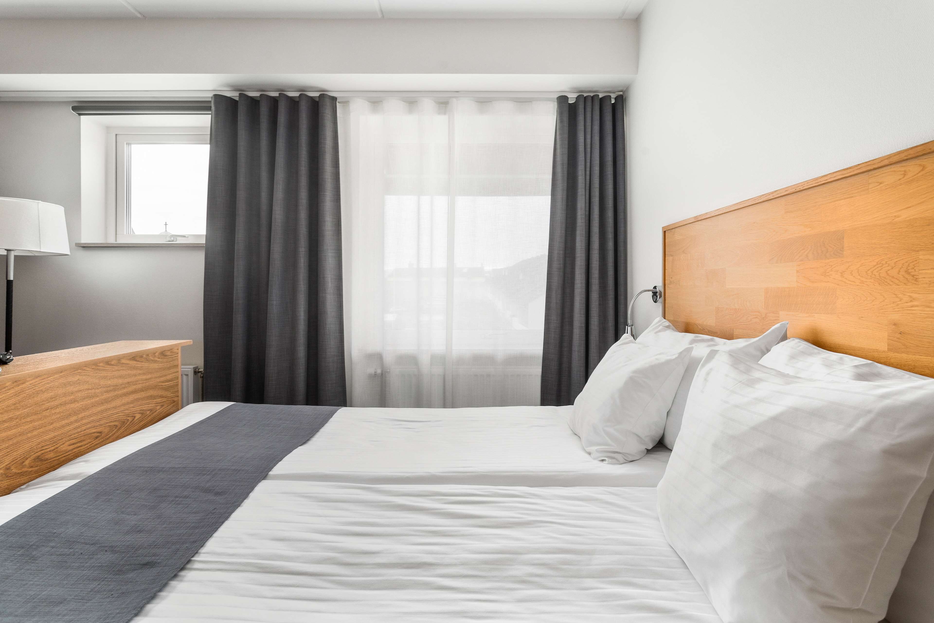 A hotel room with a bed, a wooden headboard, a bedside table with a lamp, and grey curtains.