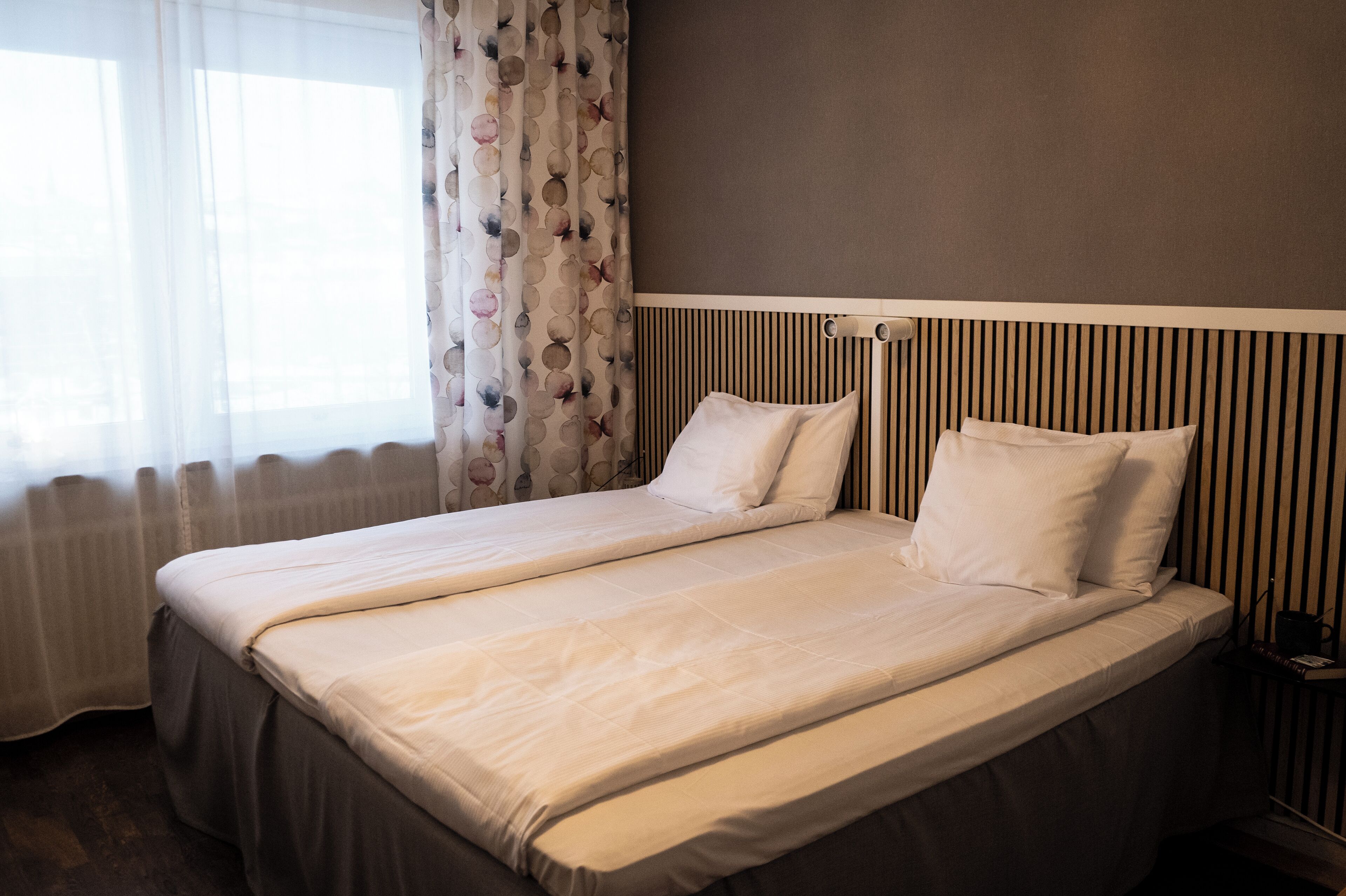 A double bed with a headboard, a bedside table with a lamp, and a window with curtains.