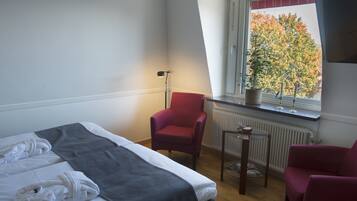 Une chambre d’hôtel avec un lit, deux chaises, une petite table, un radiateur et une fenêtre donnant sur des arbres.