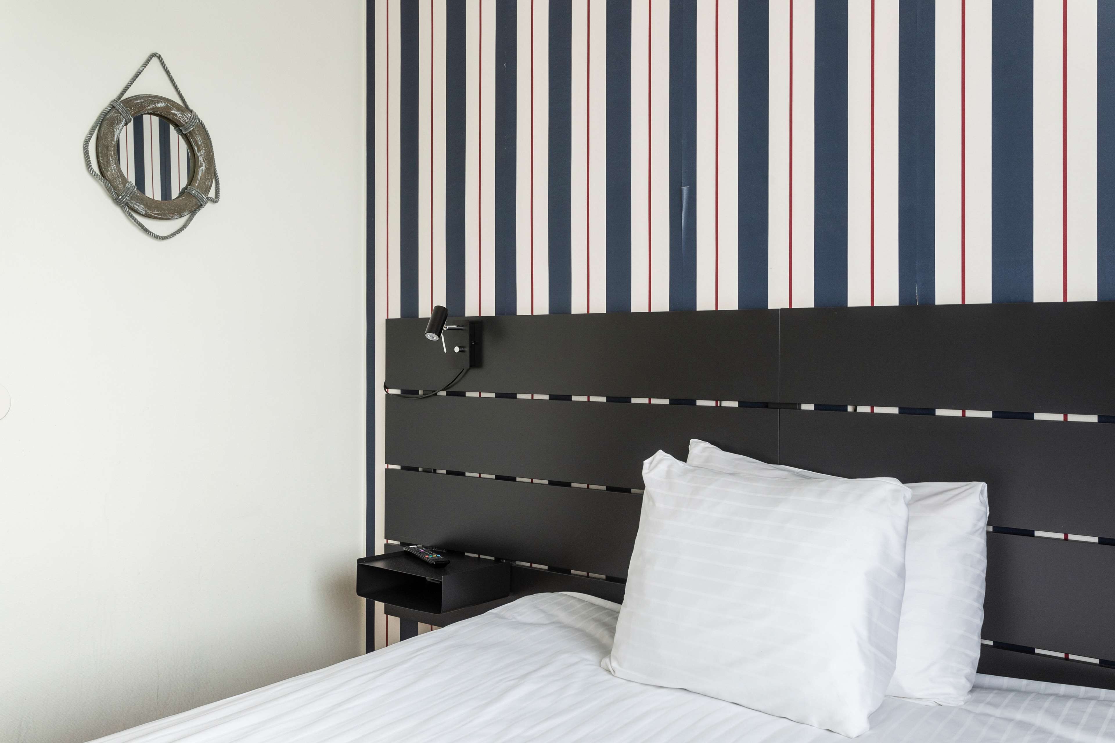 A neatly made bed with white linens, a striped wall behind it, and a small bedside table with a remote control.