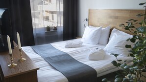 Ein ordentlich bezogenes Bett mit weißen Leinen und einer grauen Decke, ein Nachttisch mit Kerzen sowie ein Blick auf Gebäude durch ein Fenster mit dunklen Vorhängen.