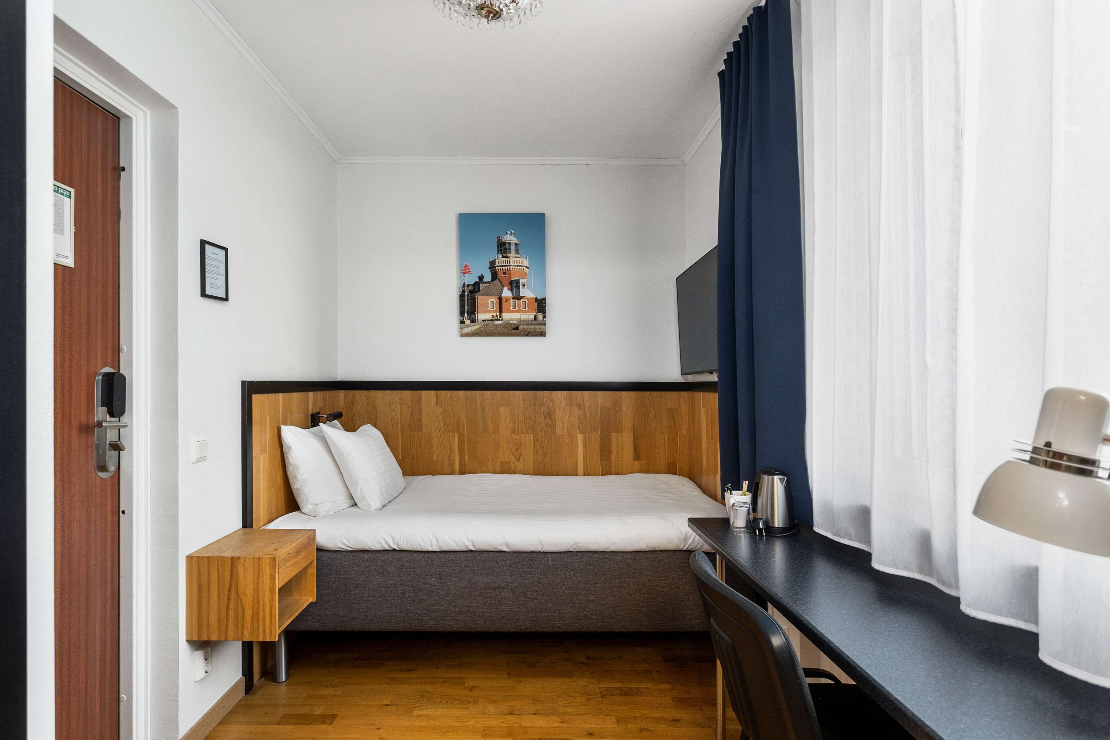 A bedroom with a wooden headboard bed, a small bedside table, a desk with a lamp, and a picture of a lighthouse on the wall.