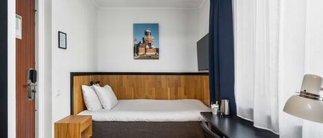 A bedroom with a wooden headboard bed, a small bedside table, a desk with a lamp, and a picture of a lighthouse on the wall.
