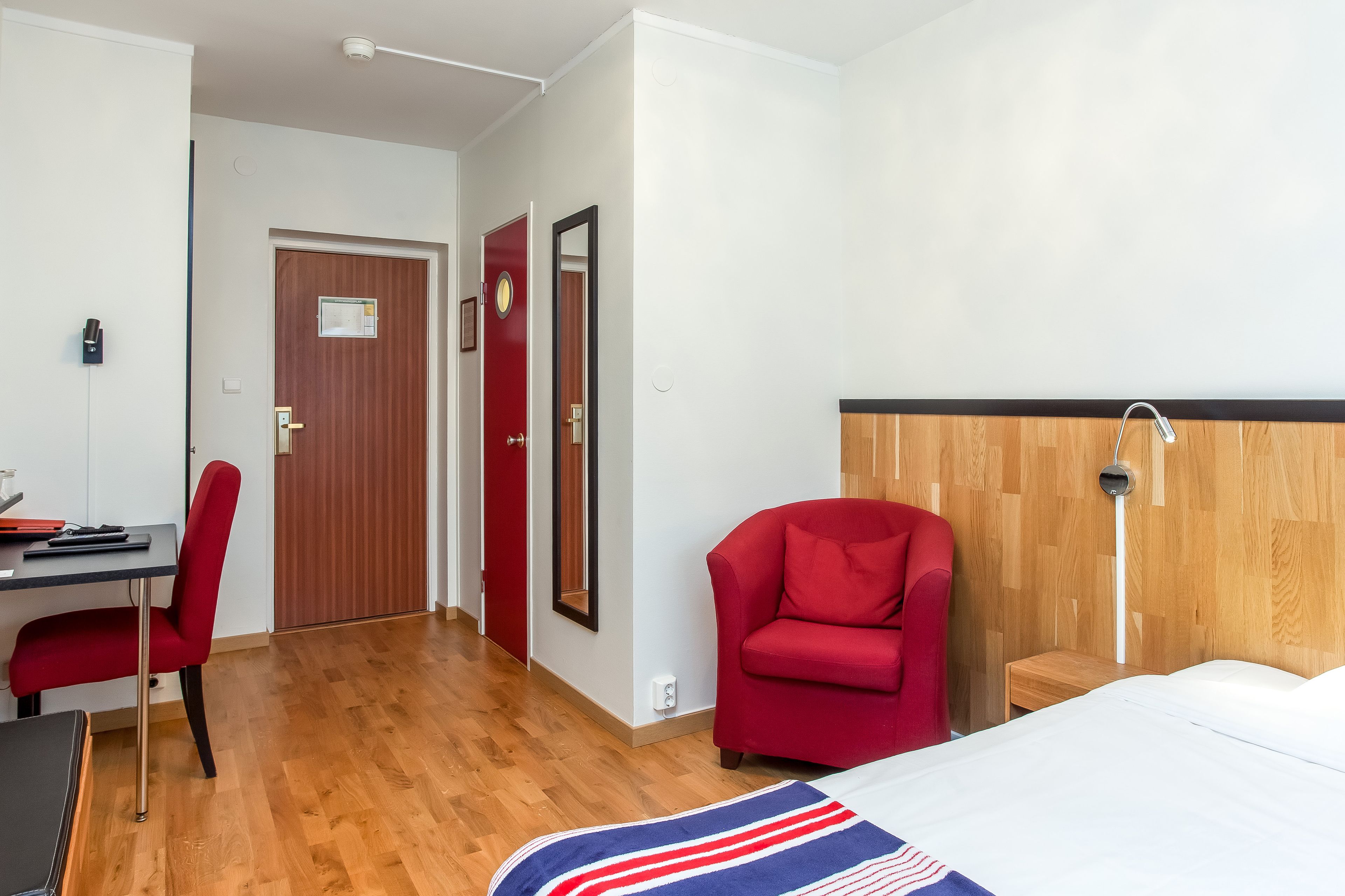 A hotel room with a wooden floor, a desk with a chair, a bed with a striped blanket, a red armchair, and a wooden headboard.