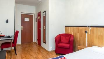 A hotel room with a wooden floor, a desk with a chair, a bed with a striped blanket, a red armchair, and a wooden headboard.