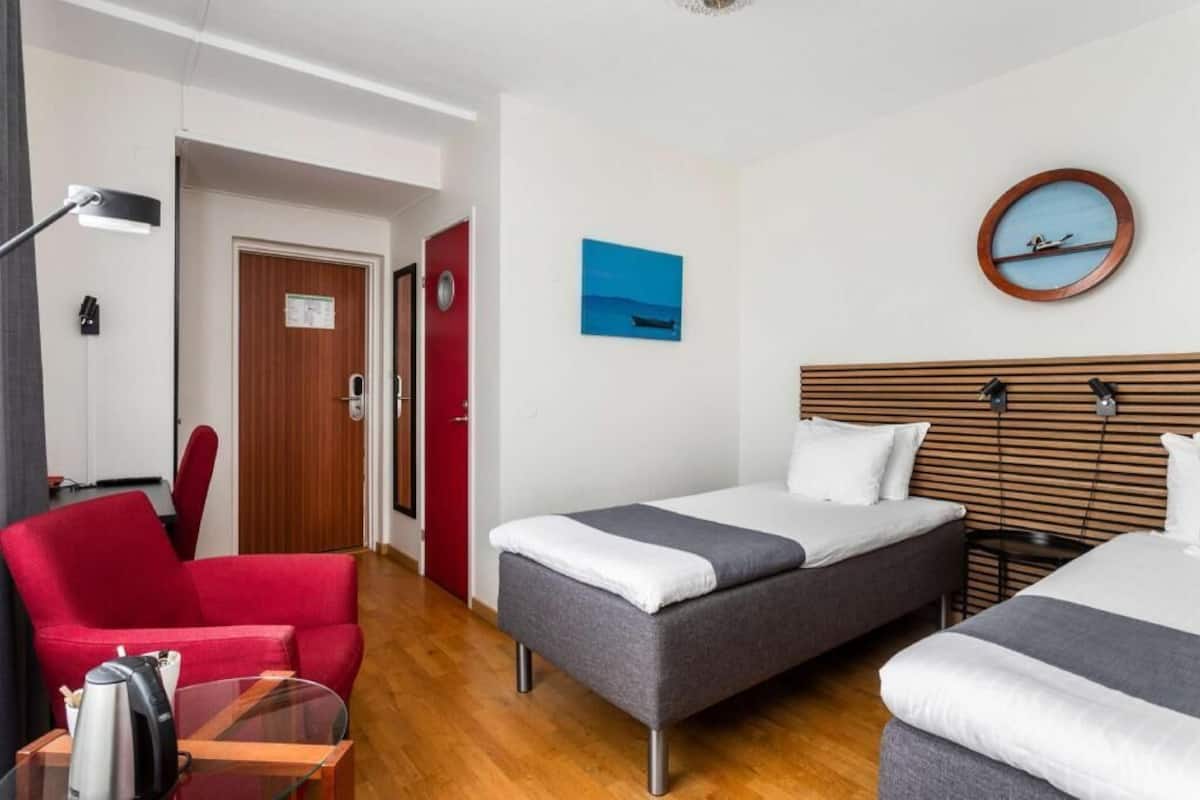 Habitación de hotel con dos camas, una silla roja, una mesita con una tetera, una lámpara y un reloj de pared.