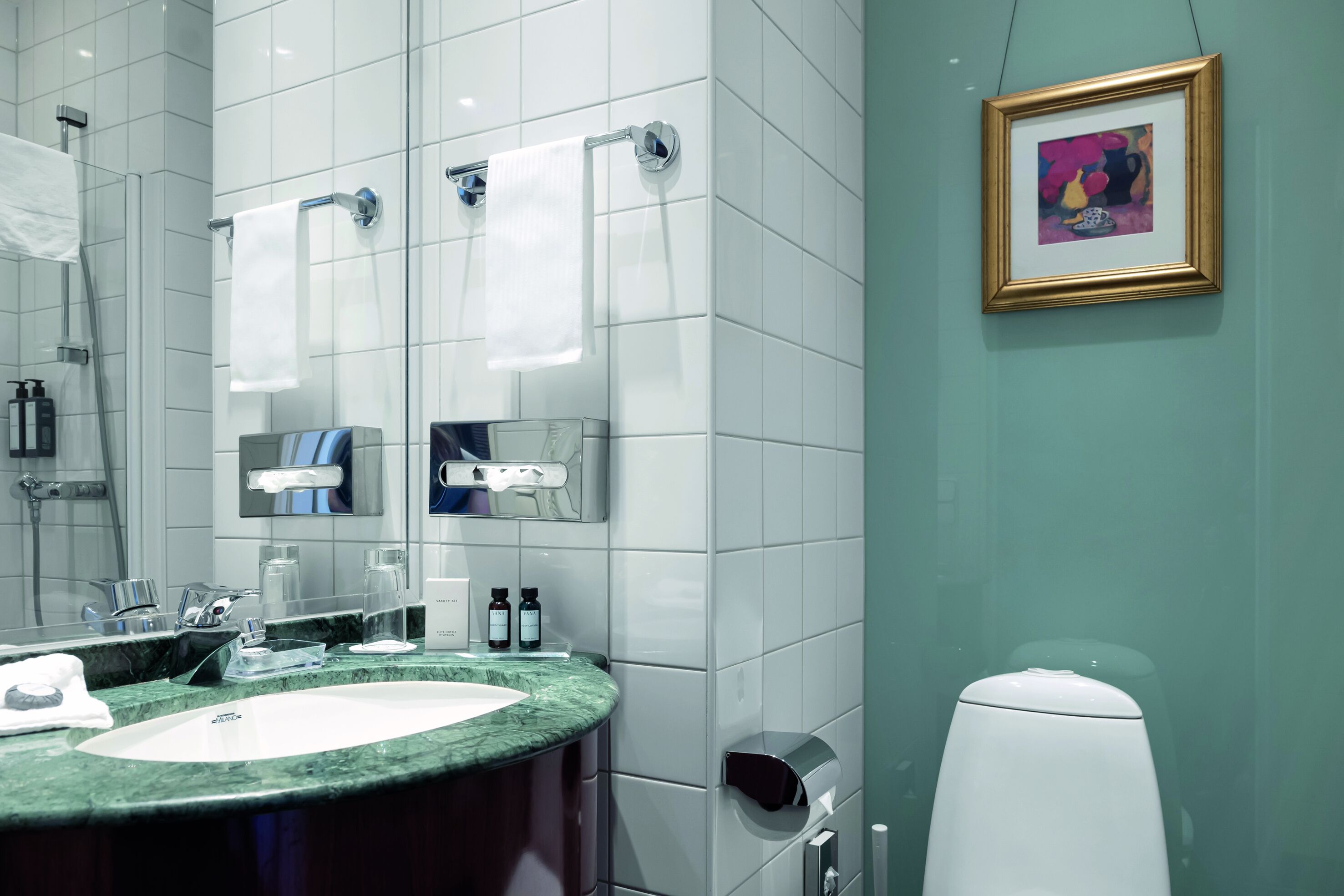 Un baño moderno con un lavabo de mármol, dos barras para toallas, un secador de manos y una obra de arte enmarcada en la pared.