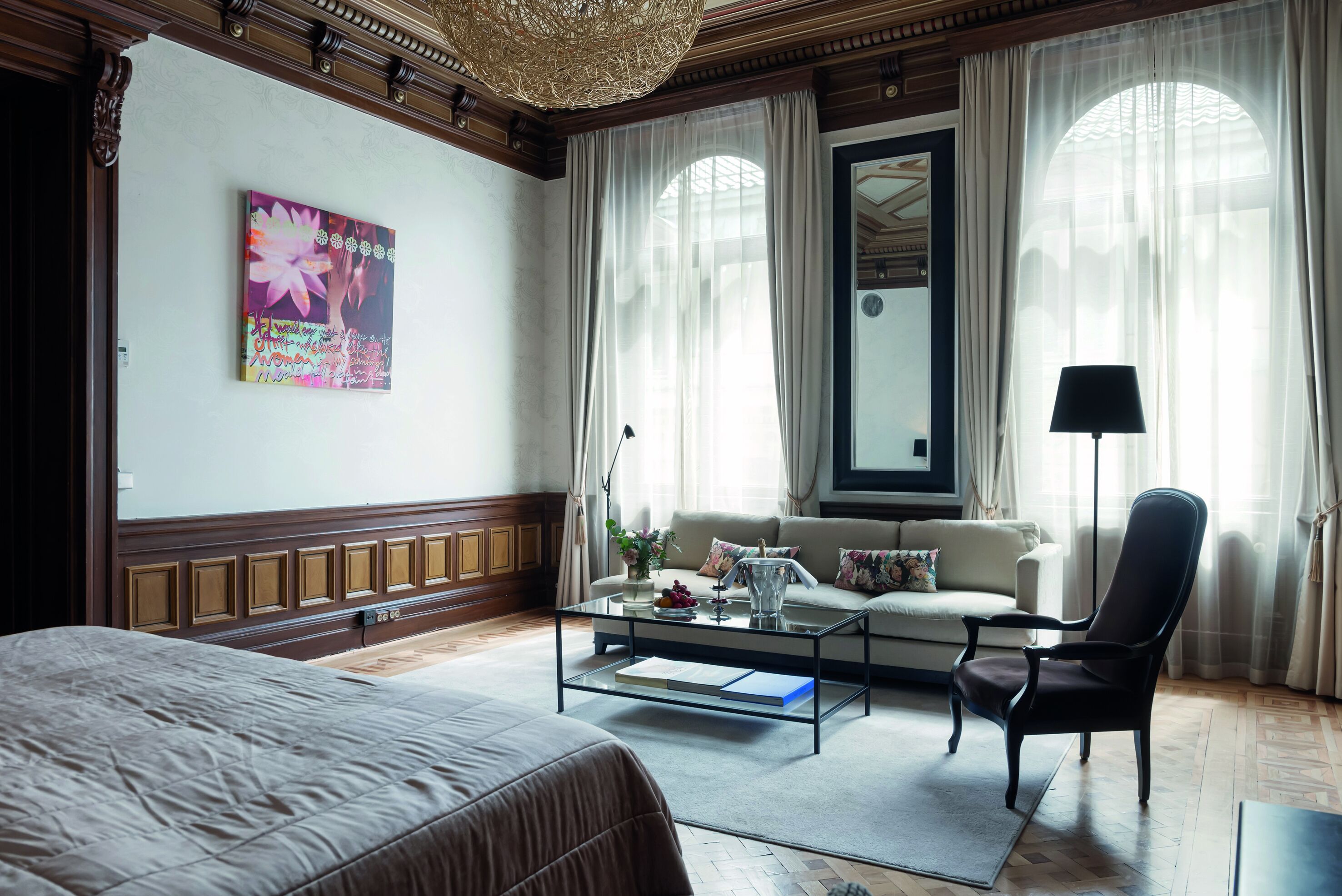 Habitación de hotel con una cama grande, un sofá, una mesa de centro y una silla. Hay dos ventanas con cortinas translúcidas y un espejo grande sobre el sofá.