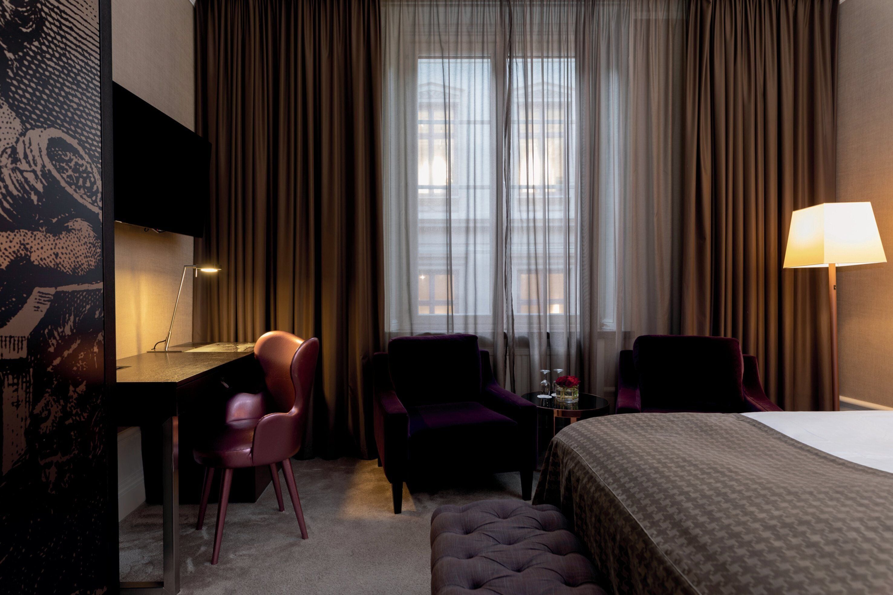 Una habitación de hotel con cama, dos sillones, un escritorio, televisor y lámpara.