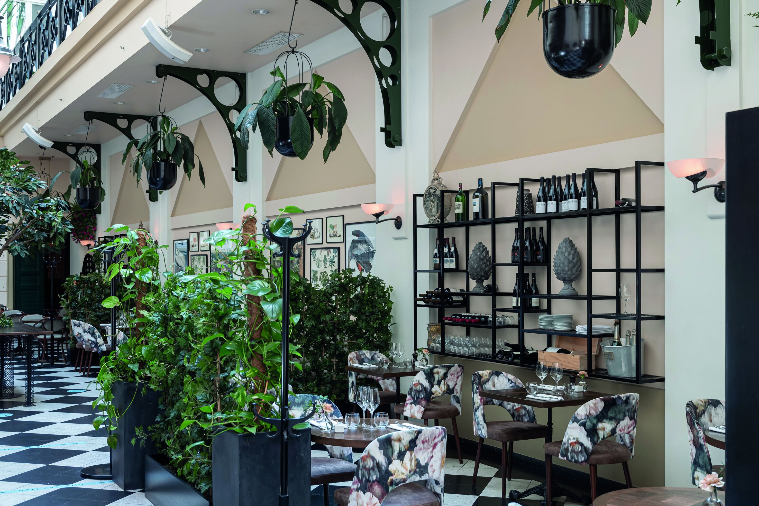 Un ristorante con pavimento a scacchi, piante sospese, sedie floreali e bottiglie di vino sugli scaffali.