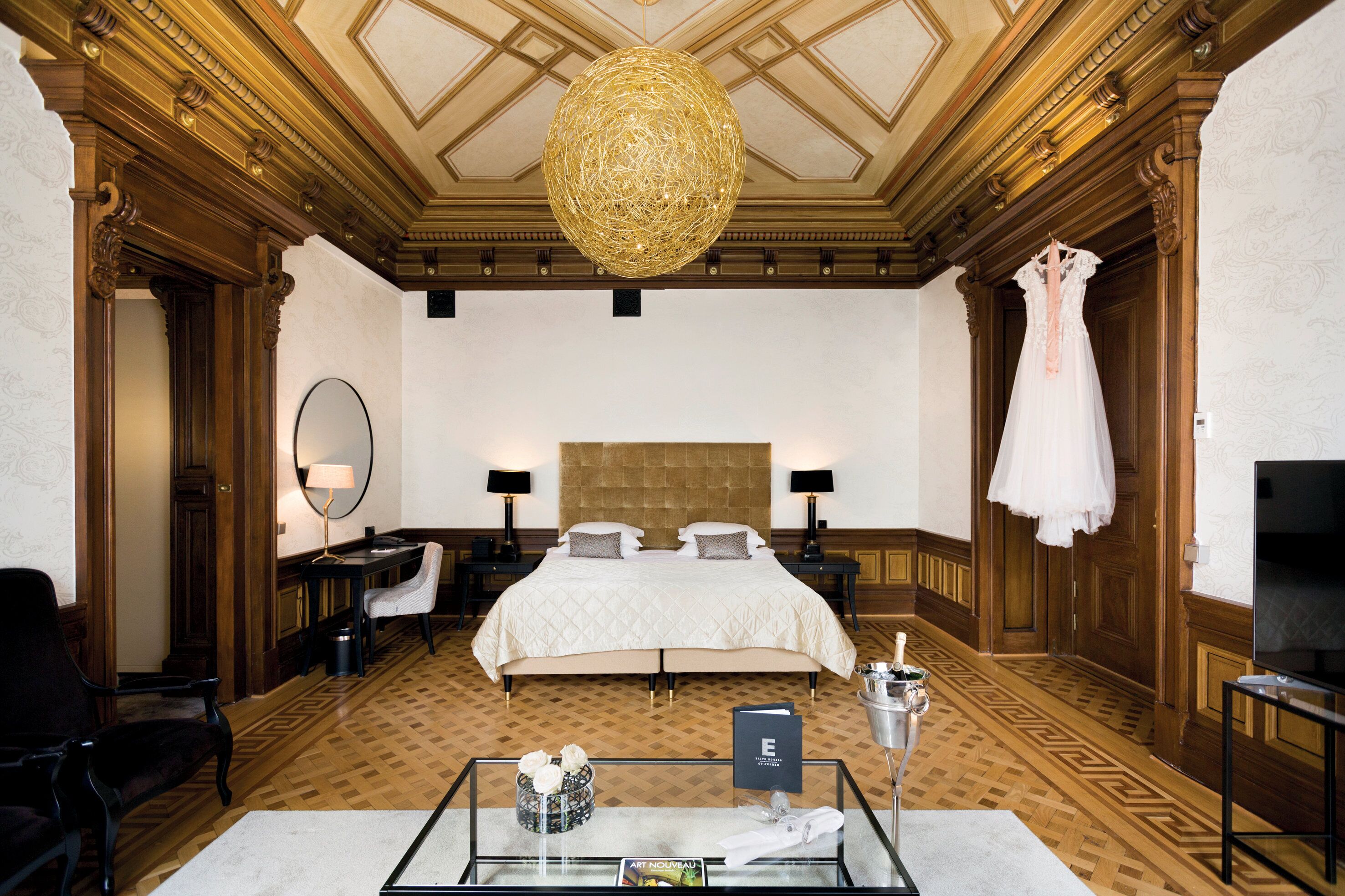 A hotel room with a large bed, a chandelier, a dress on the wall, and a desk with a lamp.