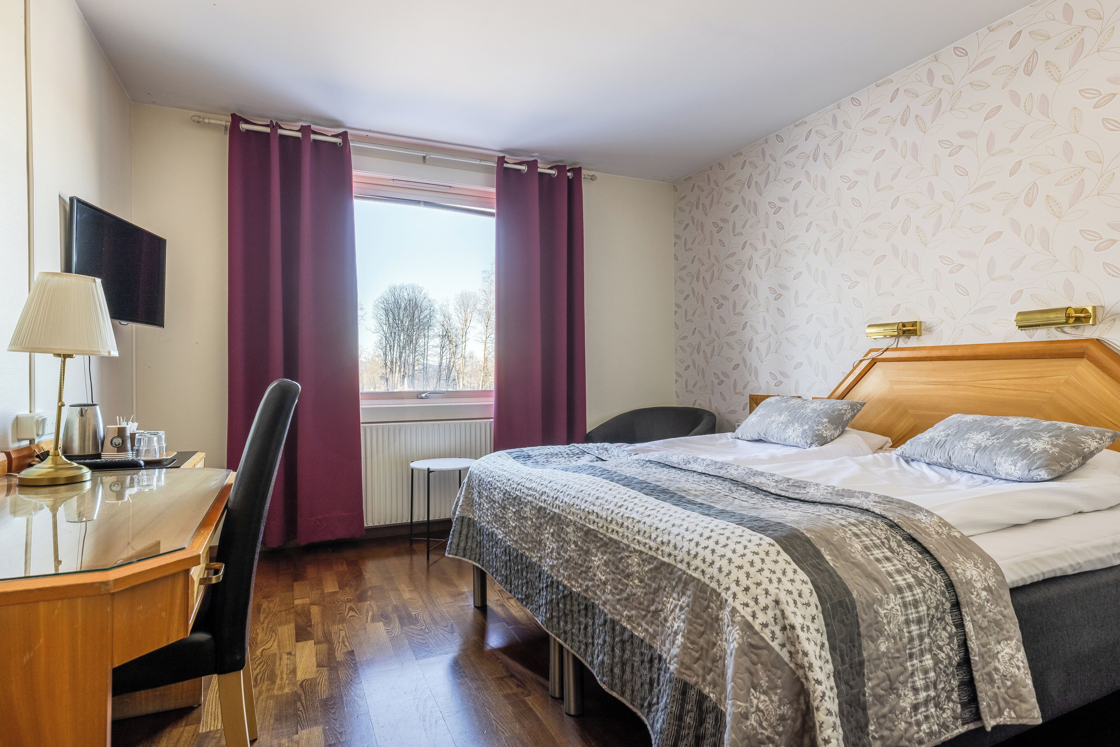 A hotel room with a wooden desk, a bed with patterned bedding, a chair, and a window with curtains.