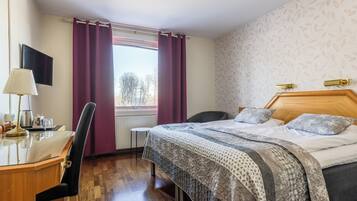A hotel room with a wooden desk, a bed with patterned bedding, a chair, and a window with curtains.
