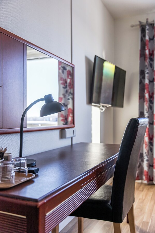 Un bureau en bois, une lampe, une chaise, un téléviseur fixé au mur et une fenêtre ornée d’un rideau à motifs floraux.