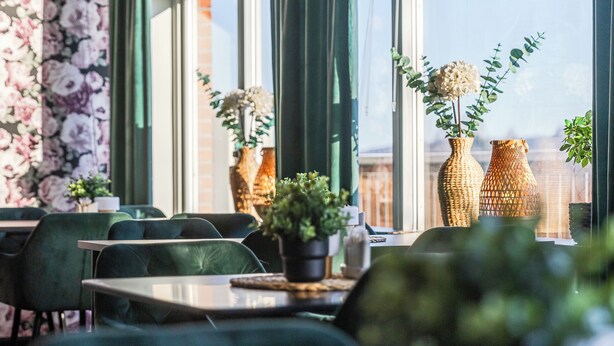 Un restaurante con sillas tapizadas de verde, mesas, jarrones de mimbre con plantas y vistas amplias a través de grandes ventanales.