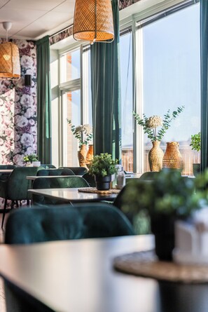 Ein Restaurant mit grünen Sitzmöbeln, Tischen, Körbchenvasen mit Pflanzen und Blick durch große Fenster.