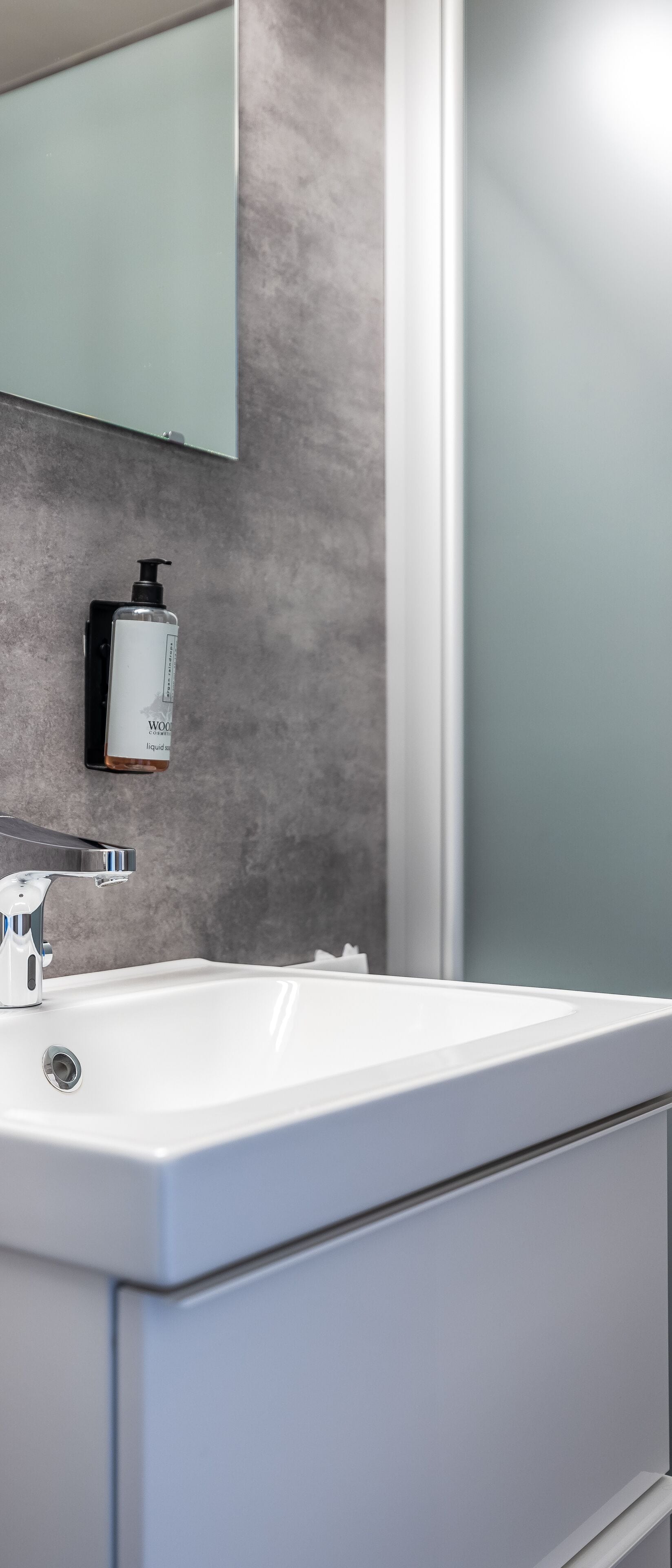 A modern bathroom with a white sink, a mirror, and a soap dispenser mounted on the wall.