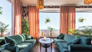 Una sala de estar moderna con dos sofás verdes, una mesa de centro redonda y vistas a la ciudad a través de ventanas grandes con cortinas rosadas.