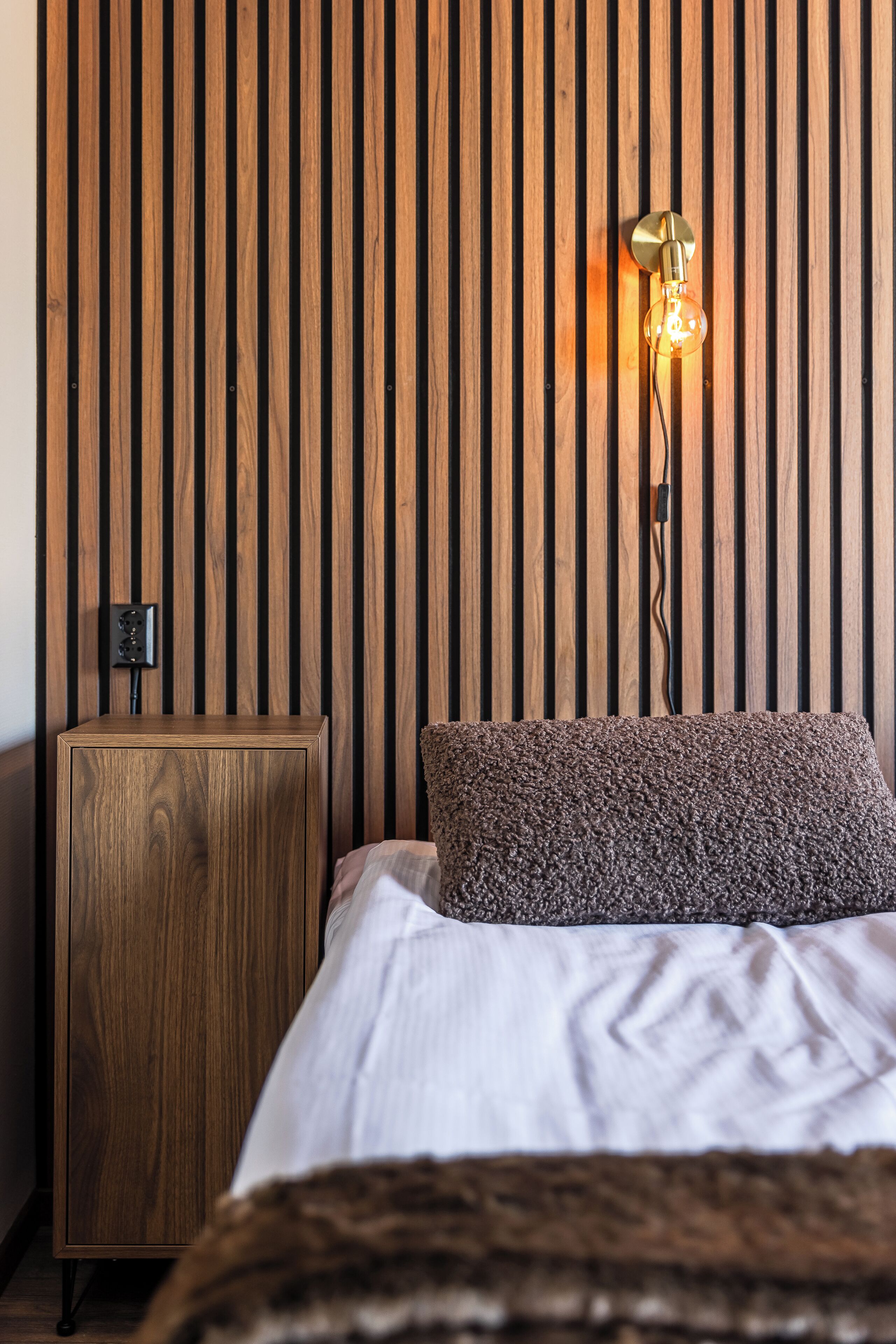 A bedroom with a wooden slat wall, a bedside table, a lamp, and a bed with a white sheet and a brown pillow.