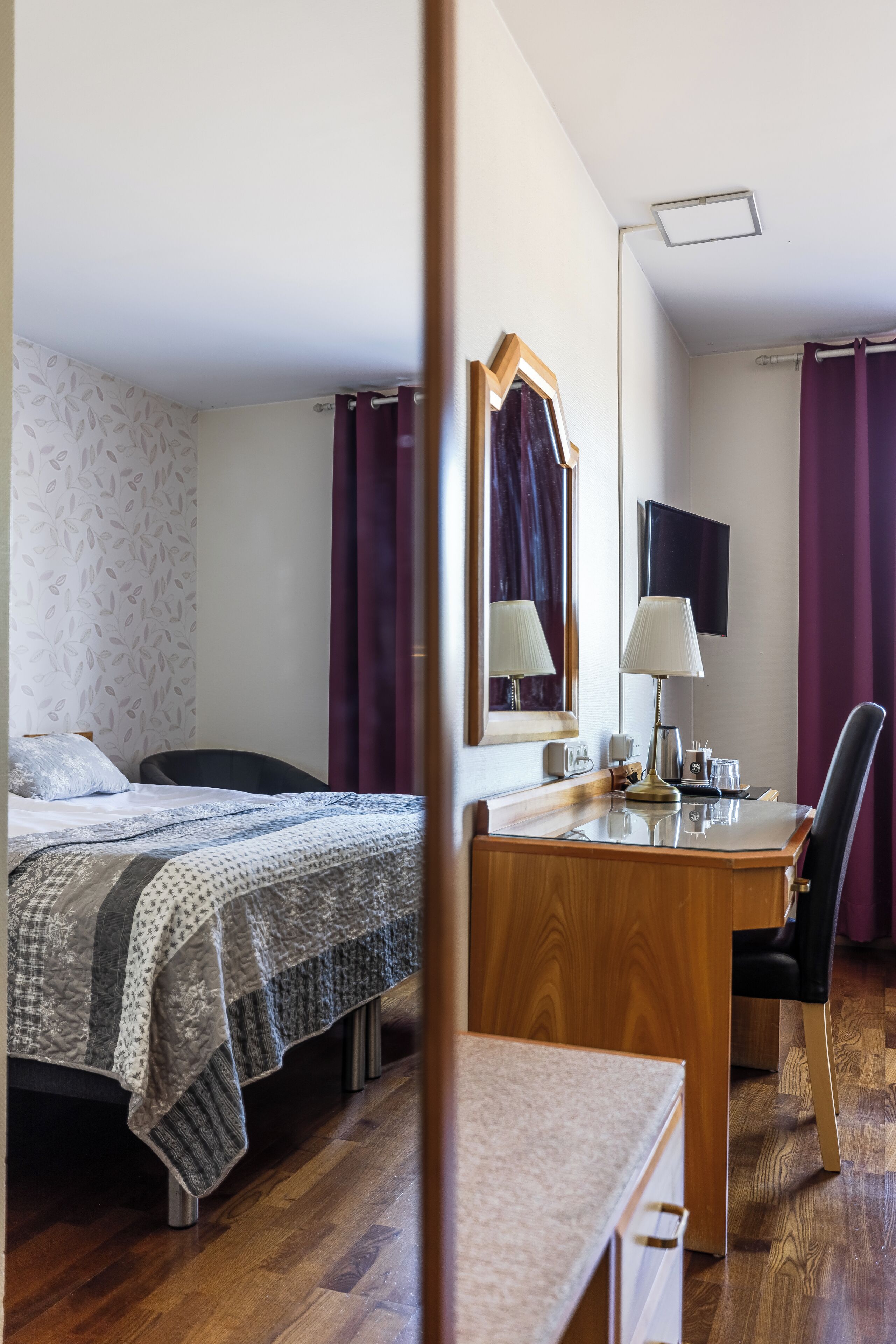Ett hotellrum med en säng, ett skrivbord, en stol, en spegel, en tv och ett fönster med gardiner.