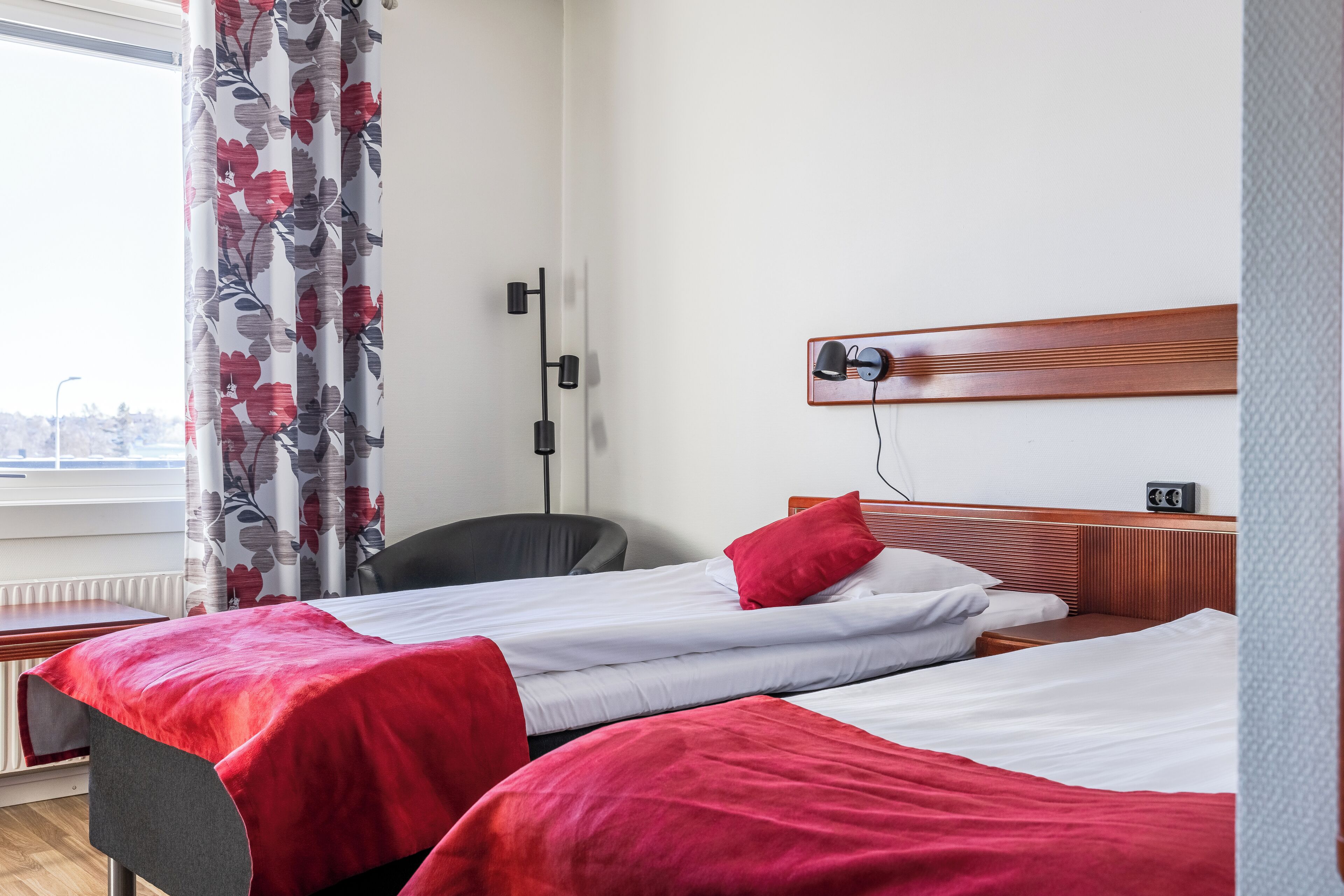 Habitación de hotel con dos camas, un escritorio, una silla, un radiador y una ventana con cortinas.