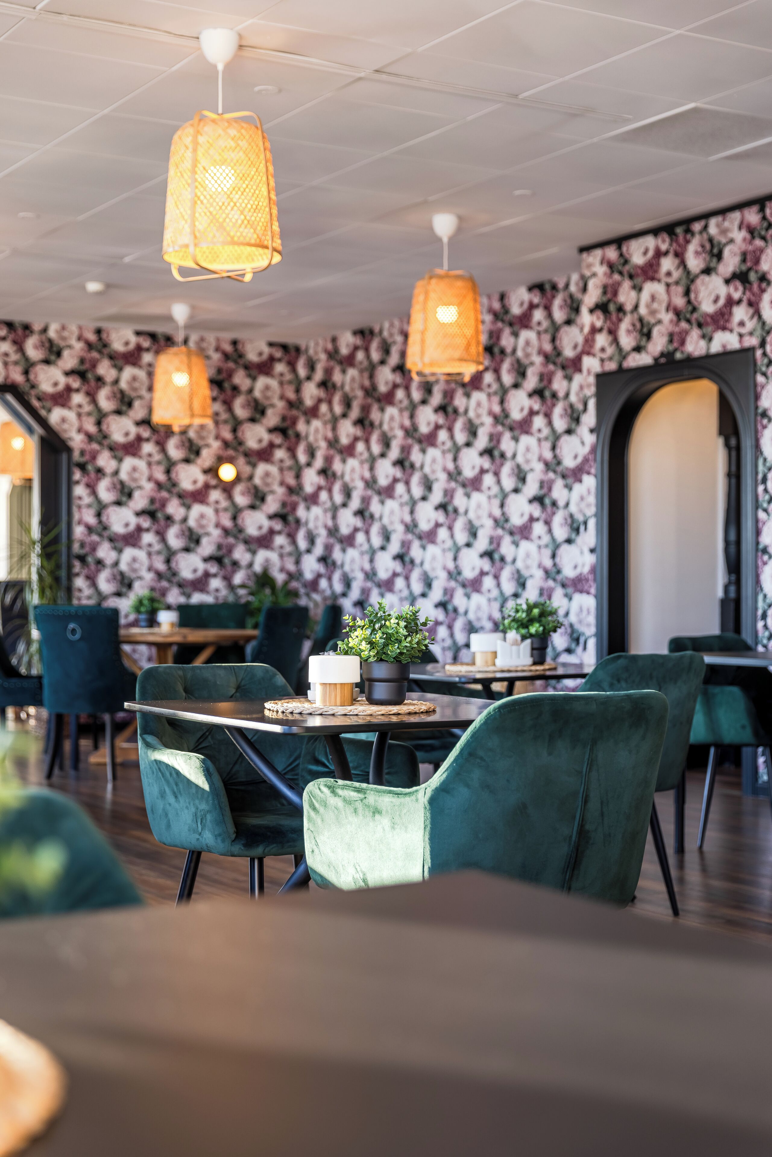 A restaurant interior with floral wallpaper, green velvet chairs, and hanging orange lamps.