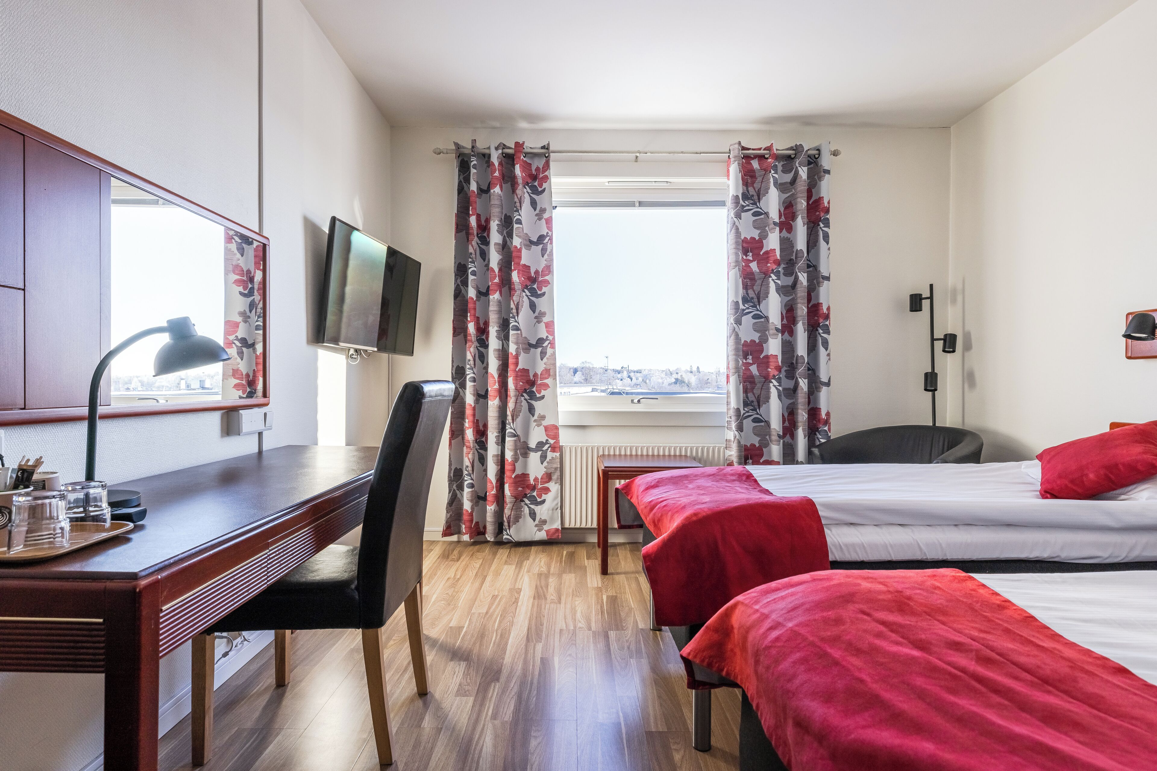 Habitación de hotel con escritorio de madera, una silla, dos camas con ropa de cama roja, un televisor montado en la pared y una ventana con cortinas florales.