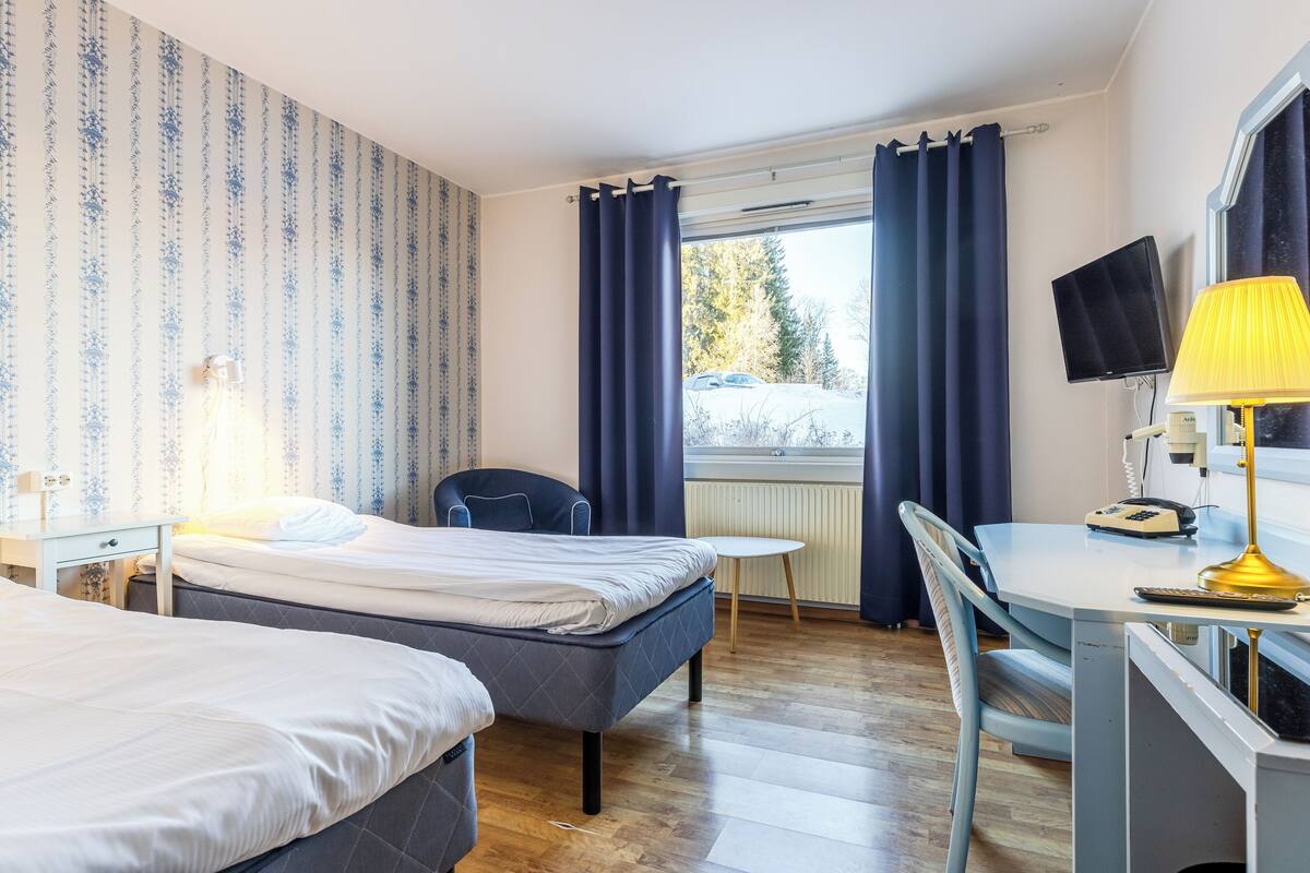 Habitación de hotel con dos camas, un escritorio con lámpara, una silla, televisor y ventana con cortinas.
