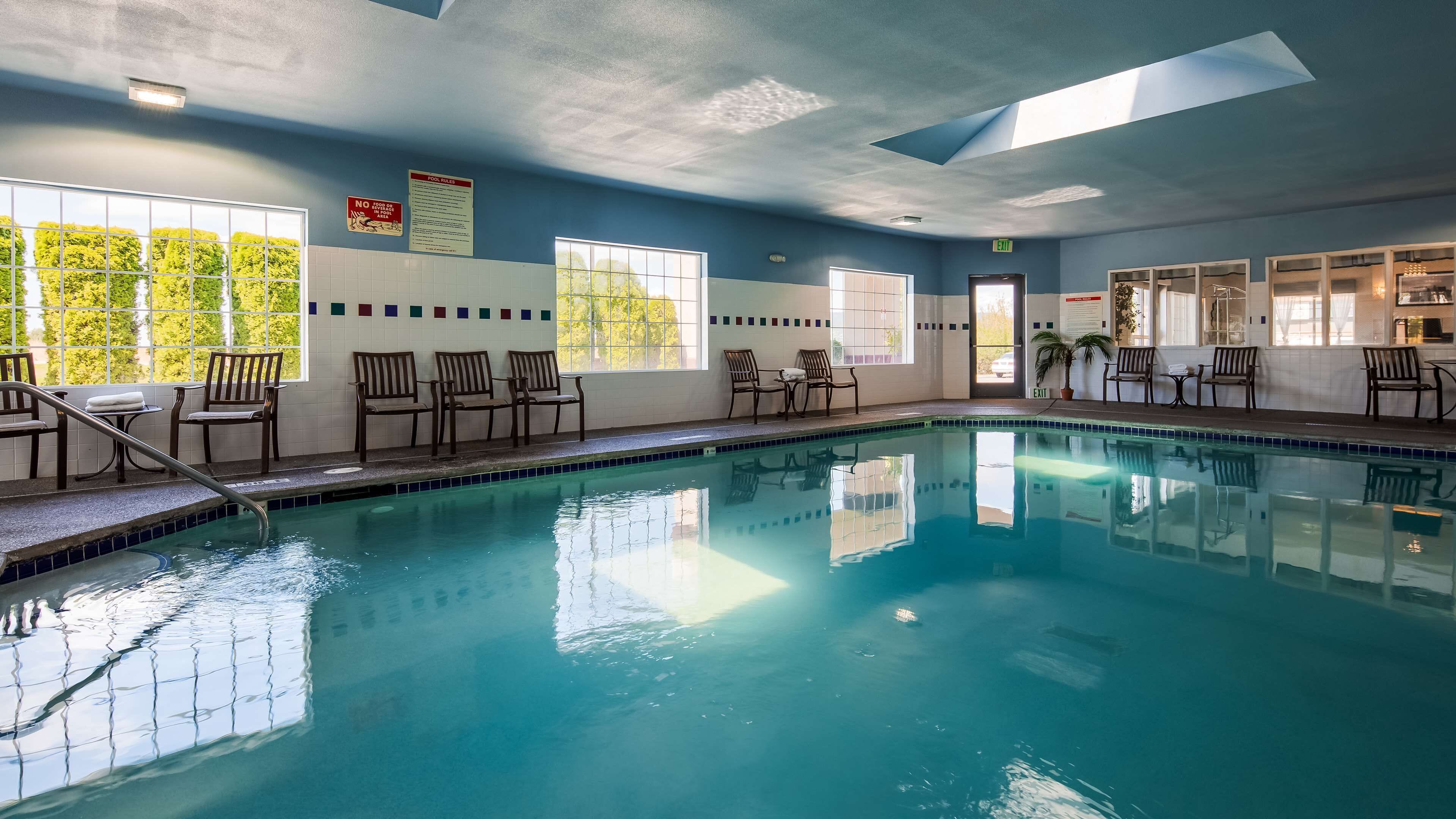An indoor swimming pool with clear blue water, surrounded by wooden chairs and tables, with windows allowing natural light and views of greenery outside.