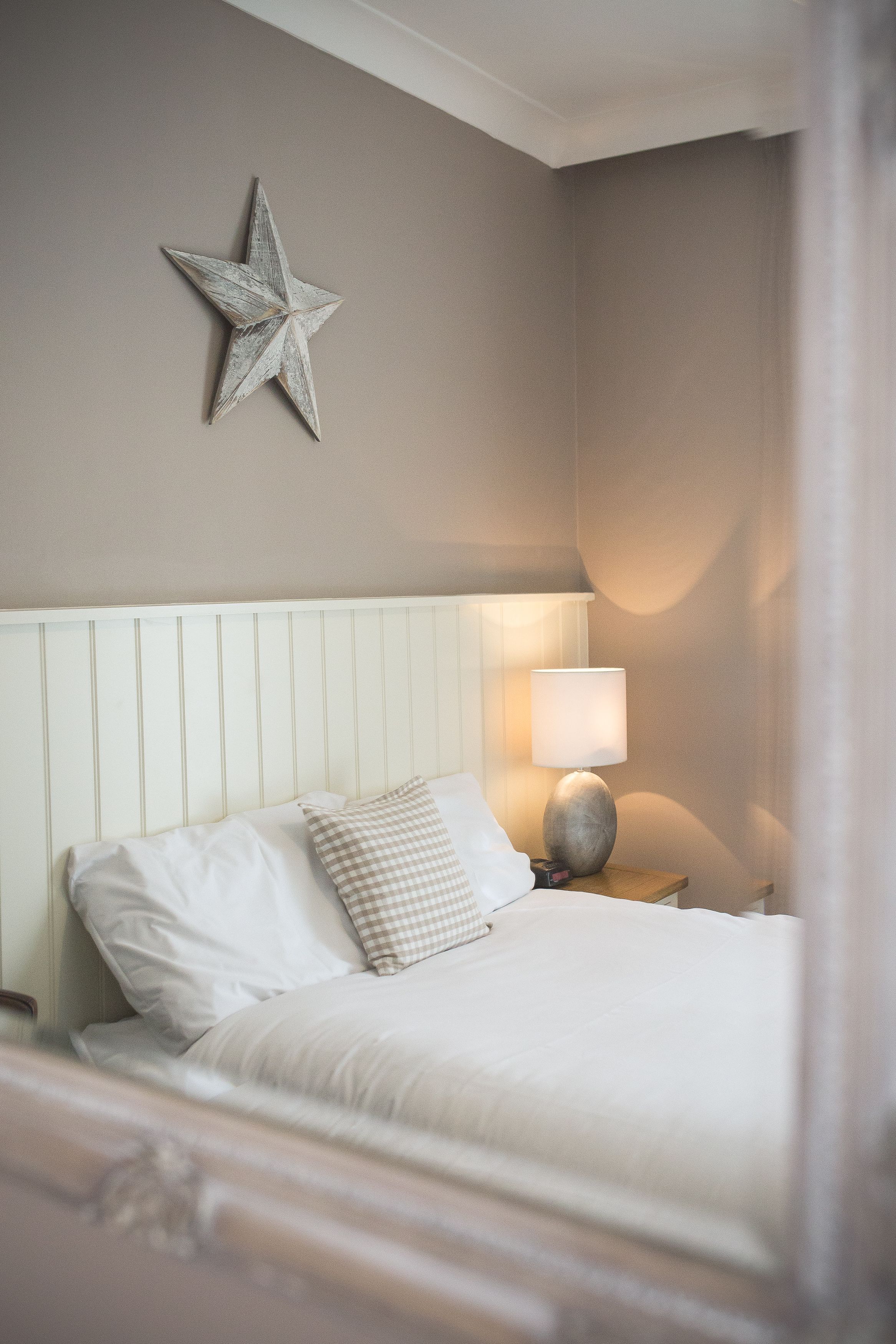 Ein ordentlich bezogenes Bett mit weißen Leinen und einem karierten Kopfkissen, eine Nachttischlampe und eine Stern-Dekoration an der Wand.