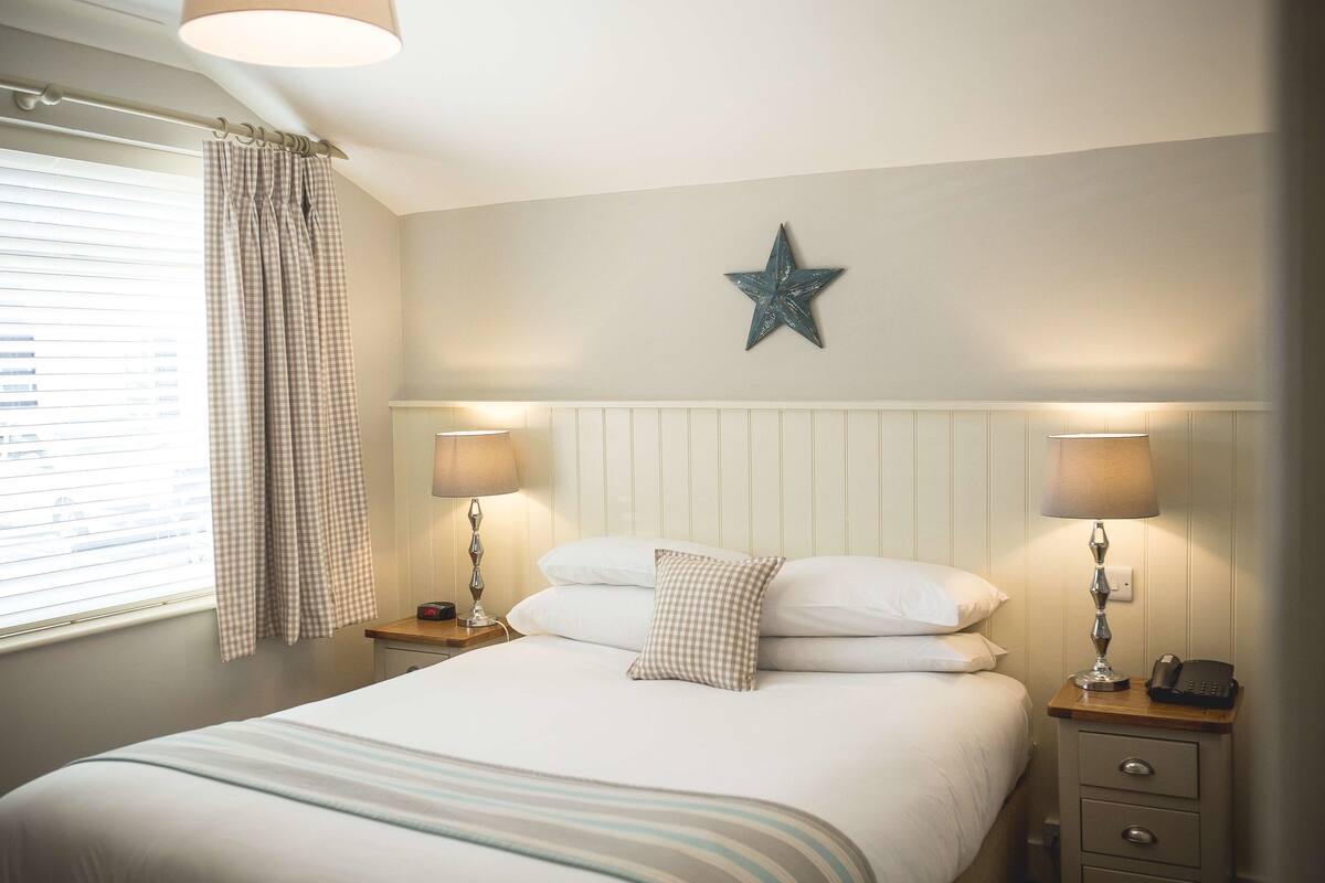 Un lit bien fait, recouvert d’une couverture à rayures, deux lampes de chevet, une décoration en forme d’étoile accrochée au mur et une fenêtre avec des rideaux.