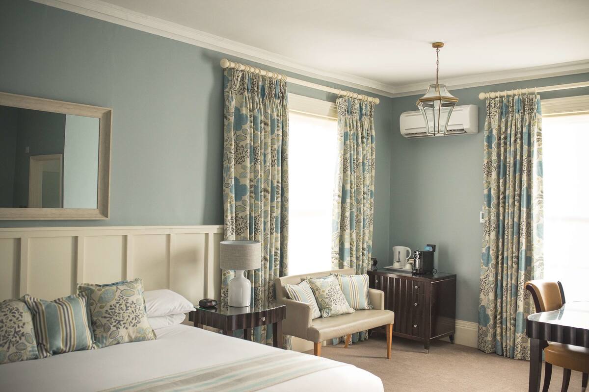 Une chambre d’hôtel avec un lit, un banc, une table et une chaise. Il y a un miroir sur le mur et un appareil de climatisation au-dessus de la fenêtre.
