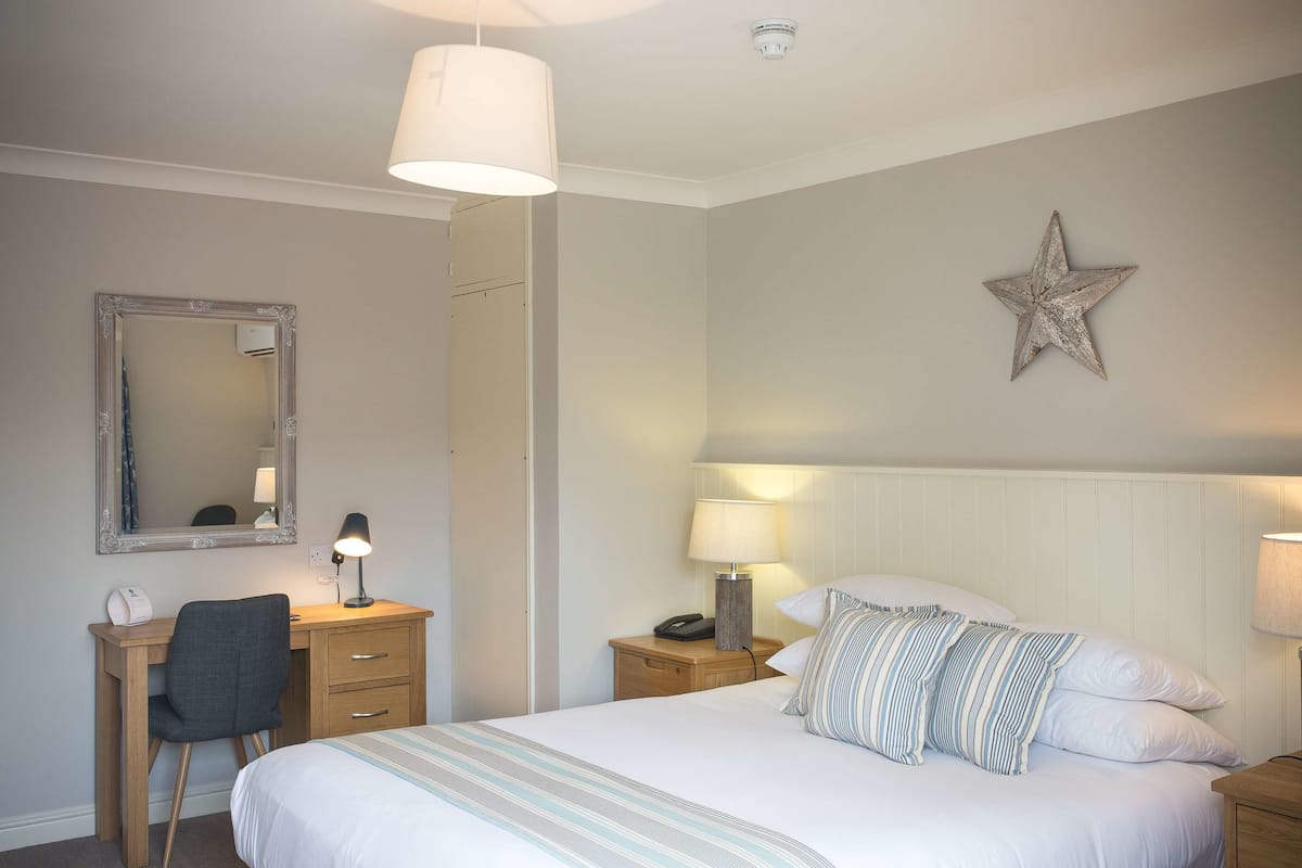 Une chambre d’hôtel bien rangée, avec un lit, un bureau, une chaise et une table de chevet avec une lampe.