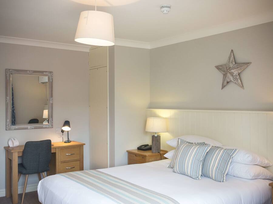 Quarto de hotel bem arrumado, com uma cama, escrivaninha, cadeira e mesa de cabeceira com abajur.