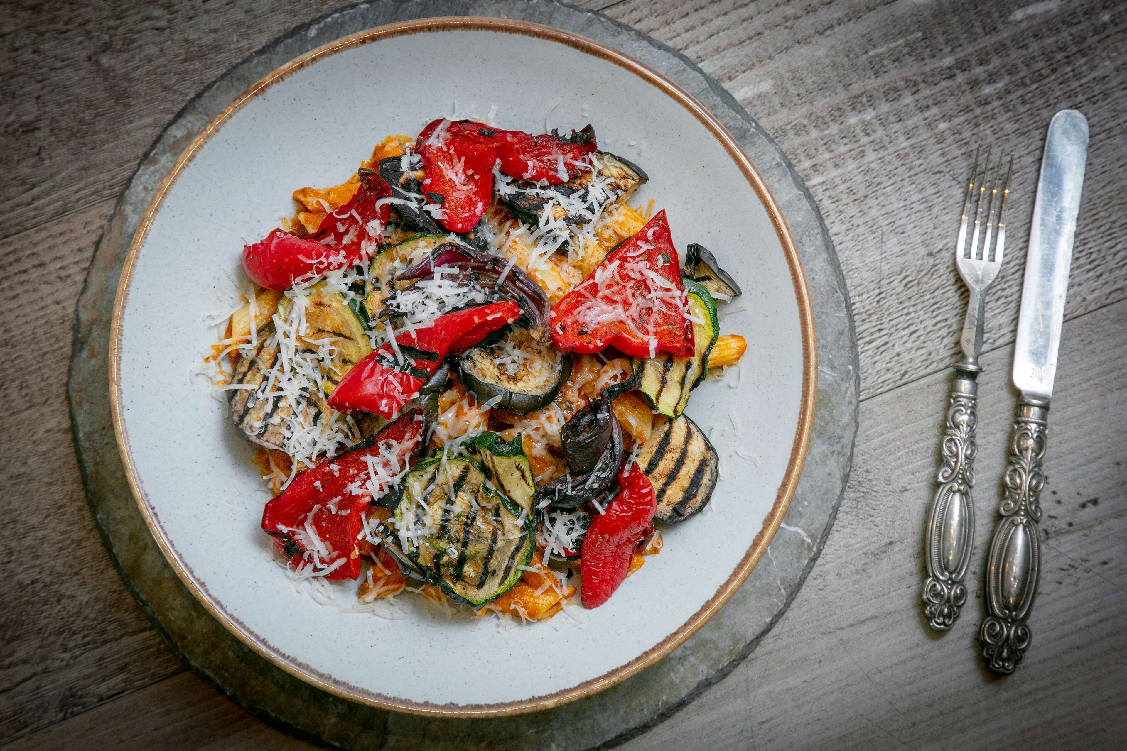 Een bord pasta met gegrilde groenten en kaas, geserveerd op een rond bord.