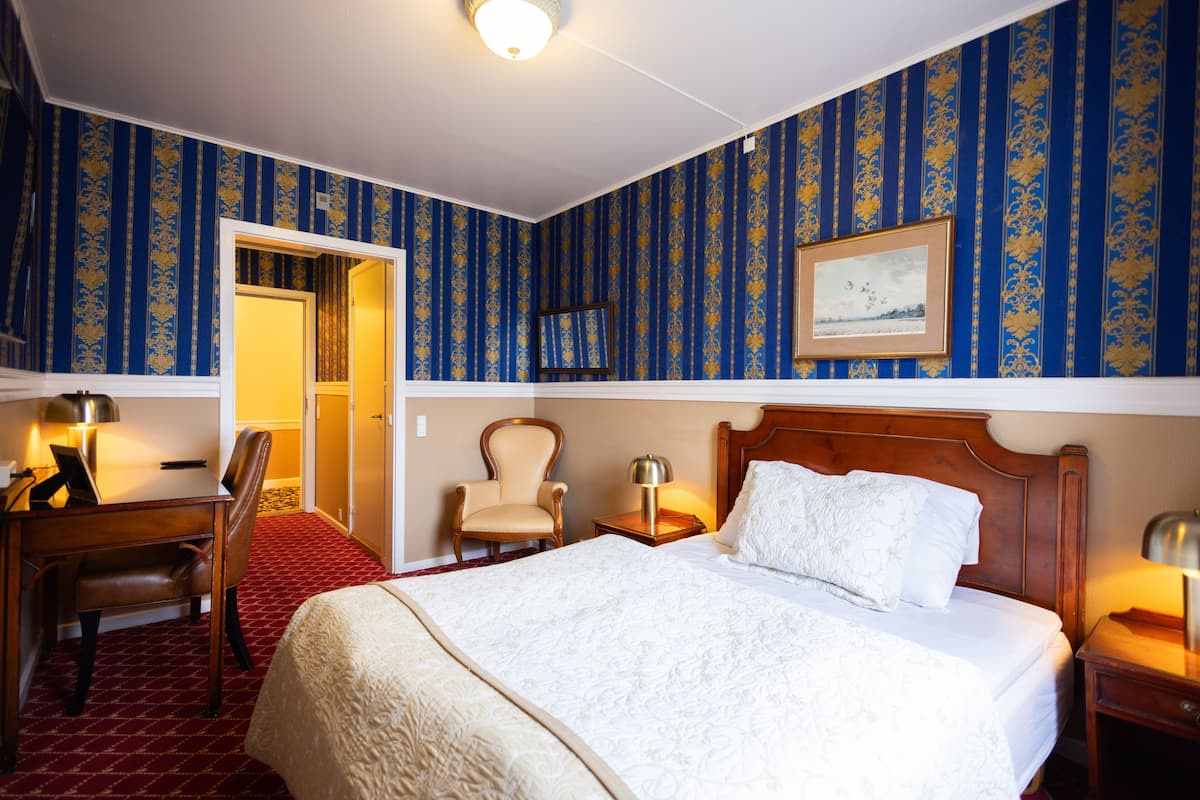 A hotel room with a large bed, a desk with a chair, a small table with a lamp, and a chair.