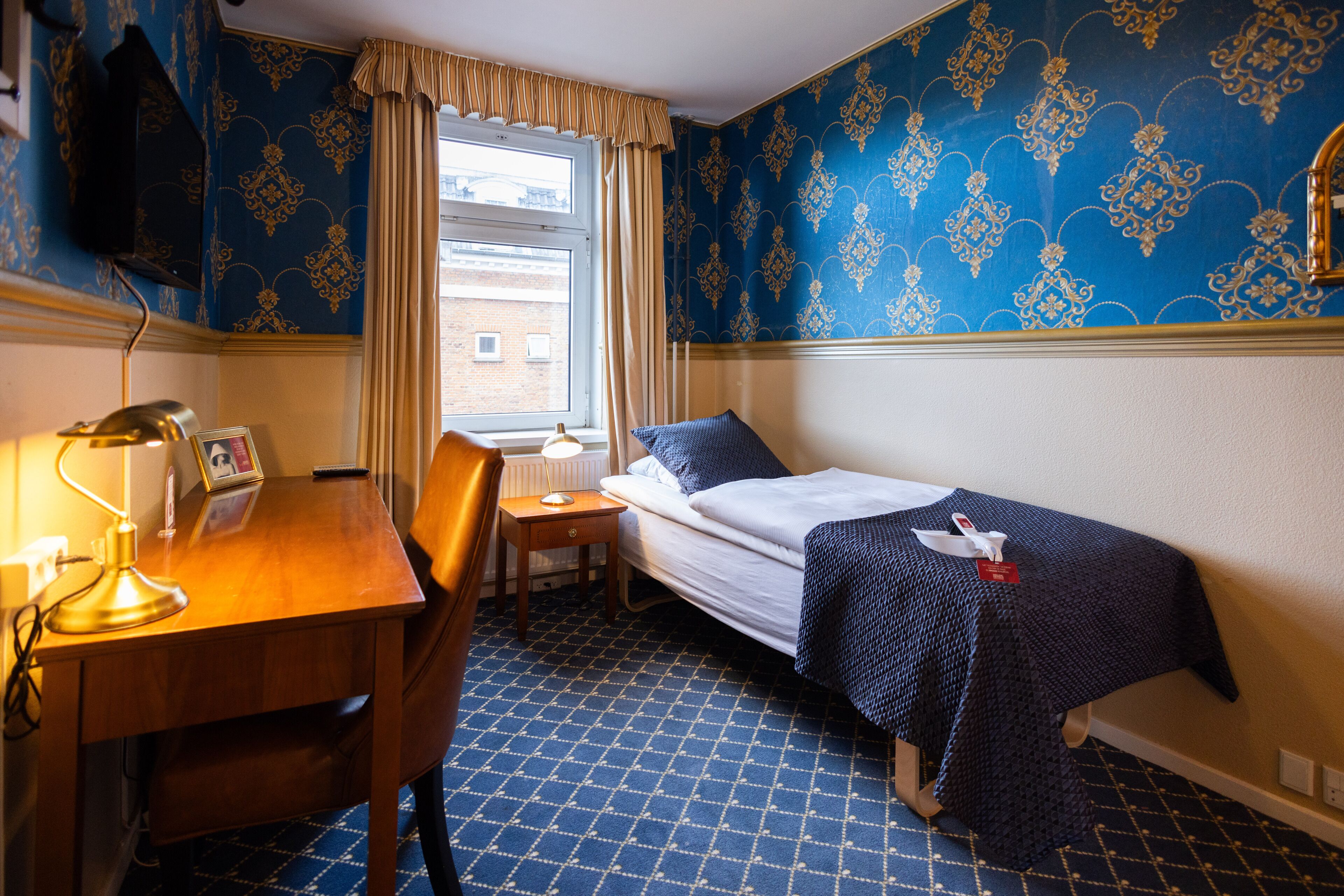 Une chambre d’hôtel avec un lit simple, un bureau avec une lampe, une chaise, une fenêtre avec des rideaux et un papier peint à motifs.