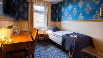 A hotel room with a single bed, a desk with a lamp, a chair, a window with curtains, and patterned wallpaper.