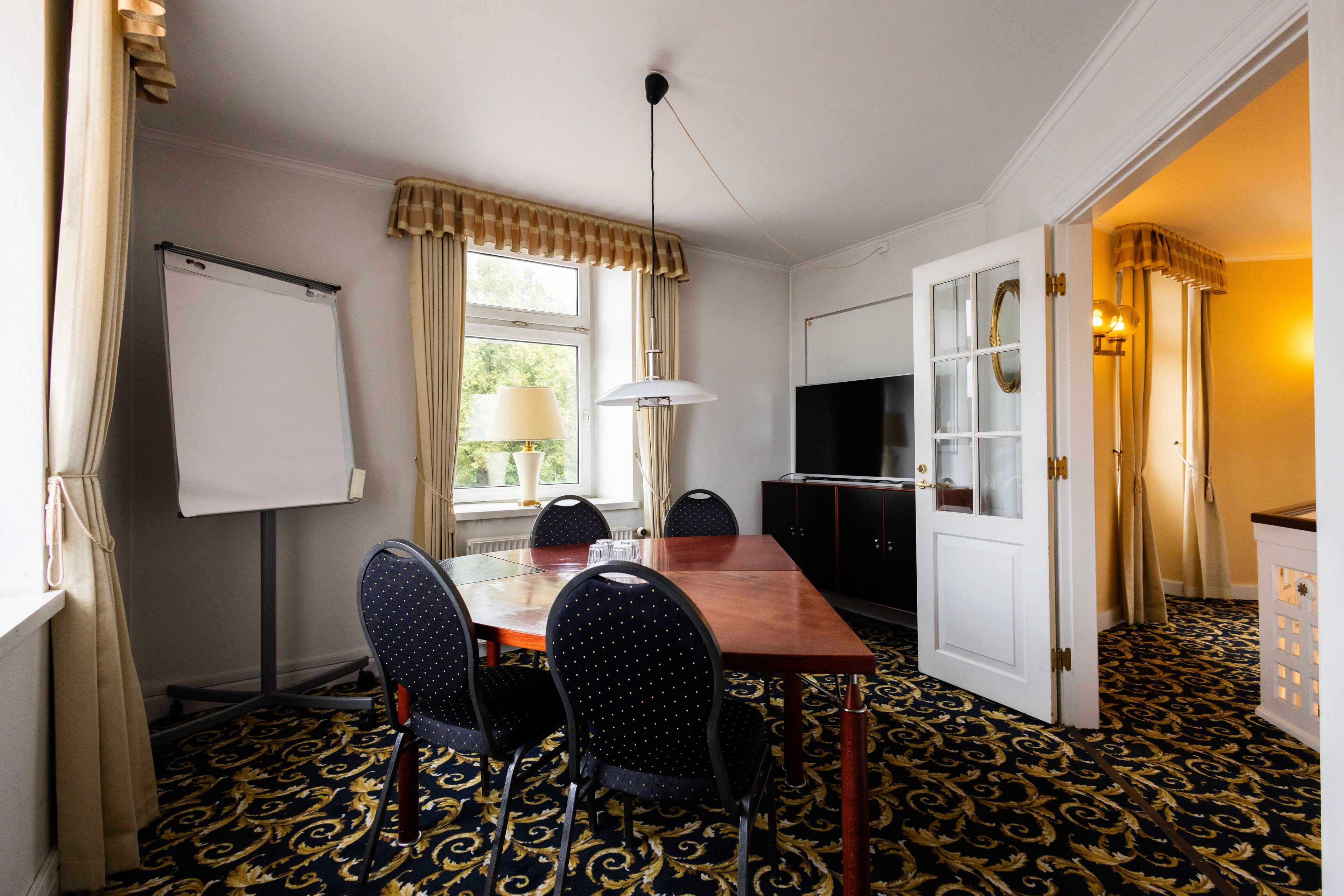 A hotel room with a dining area featuring a wooden table, four chairs, a whiteboard, a television, and a patterned carpet.