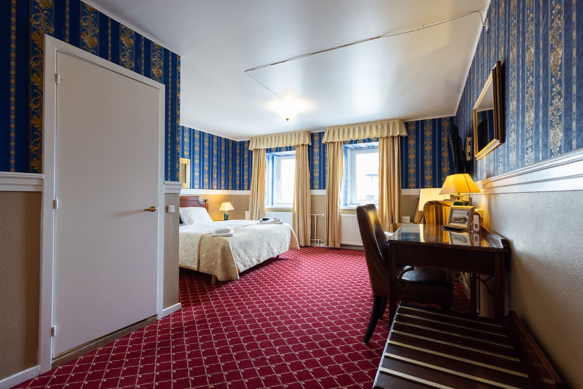 A hotel room with a red carpet, a bed with a white bedspread, a desk with a lamp, and a chair.