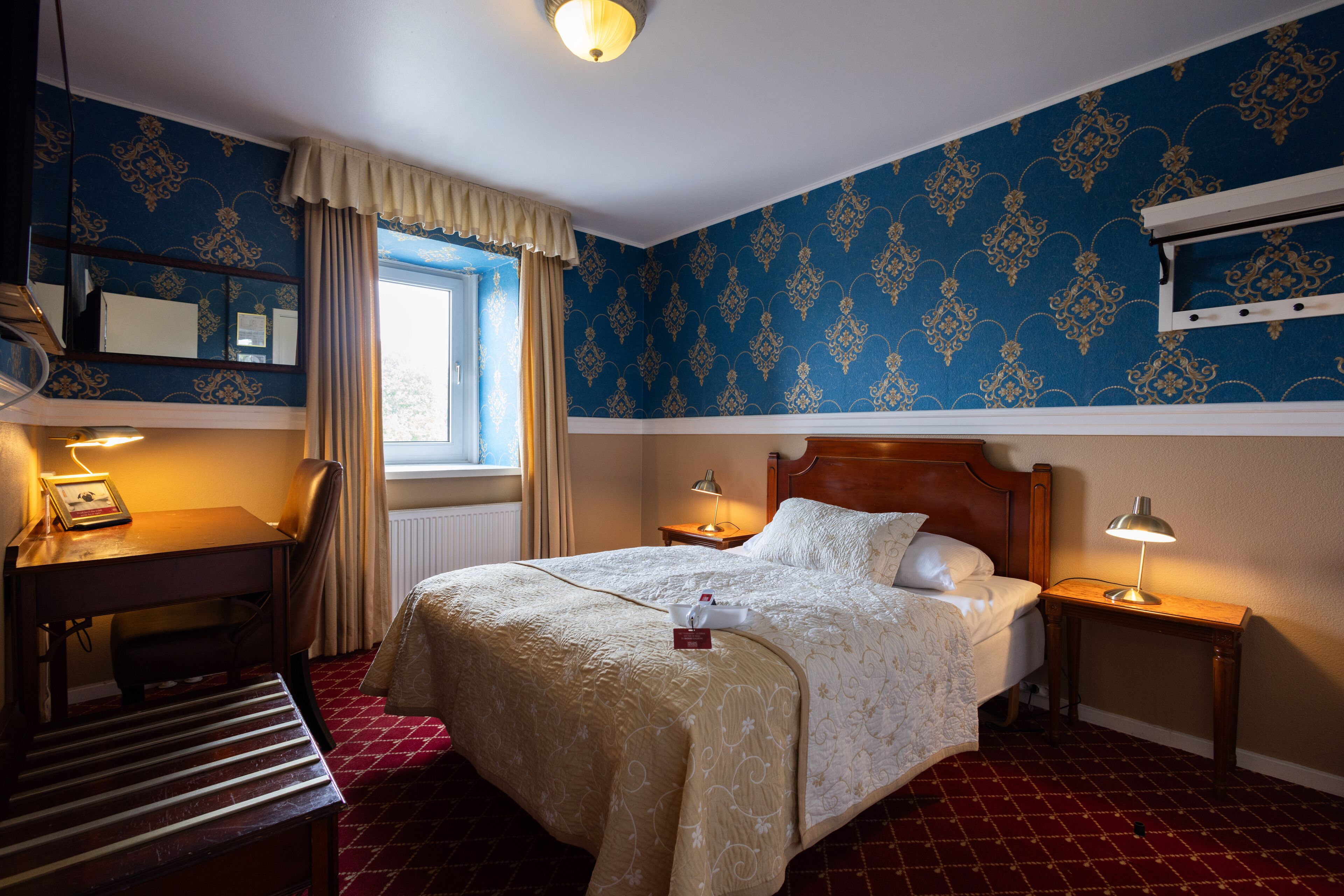 Una habitación de hotel con cama, mesitas de noche, escritorio, silla y ventana con cortinas.