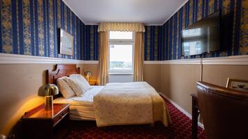 A hotel room with a bed, a nightstand, a lamp, a dresser, a television, and a window with curtains.