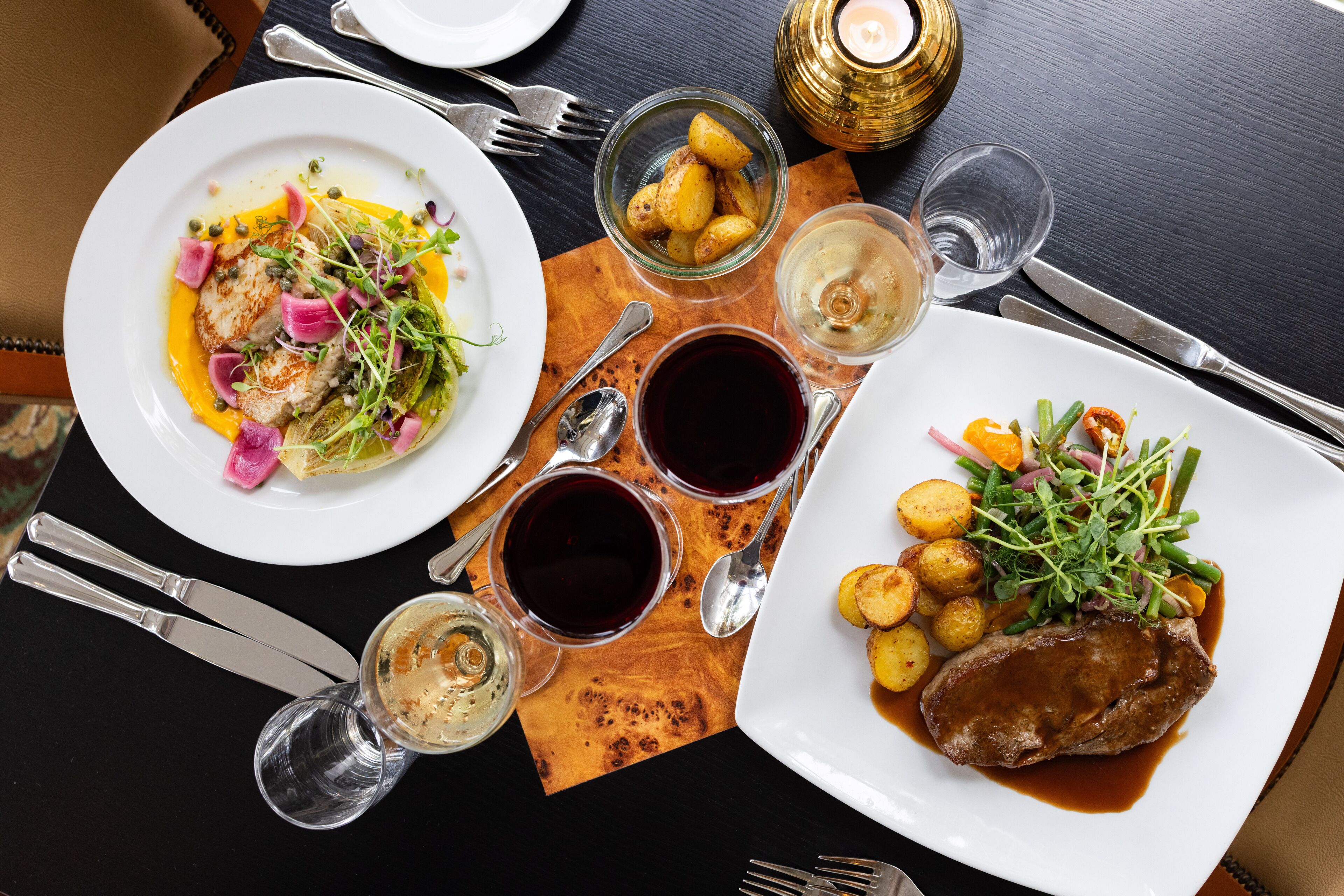 A dining table with plates of food, cutlery, and glasses of red wine.