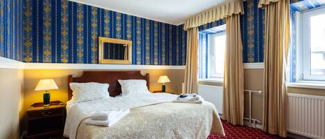 Una habitación de hotel con una cama grande, dos lámparas de noche, un cabecero de madera, una ventana con cortinas y un papel tapiz estampado.